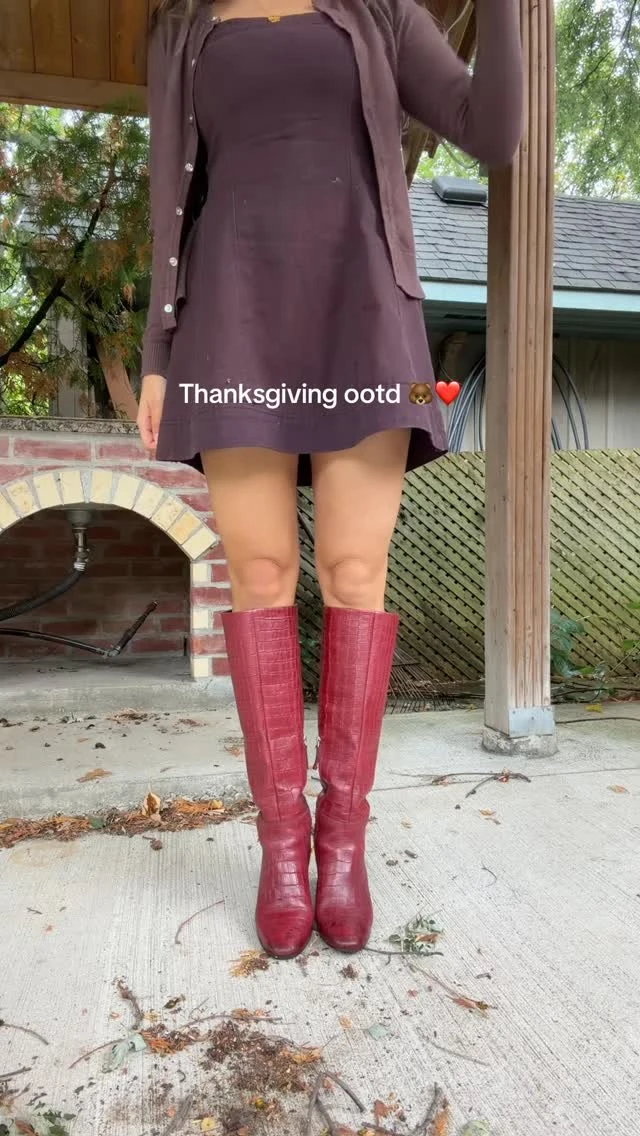 The outfit pairs a plum-colored, strapless mini dress with a matching open cardigan featuring buttons and front pockets. Tall burgundy boots reach just below the knees and have a textured, ridged finish. The person is shown outdoors, with the image cropped above the shoulders.