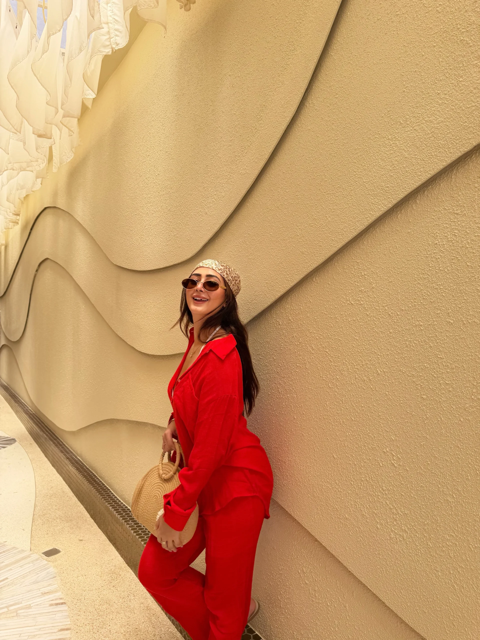 The outfit consists of a vivid red blazer worn over a matching red top and slim red trousers. The blazer has a notched collar, long sleeves and a single visible button, with a relaxed fit through the body. A light patterned headscarf covers the hair, and dark sunglasses are worn. The person carries a small rounded tan woven bag with a short handle. The visible portion of the trousers continues toward the lower edge of the image; shoes are not visible.