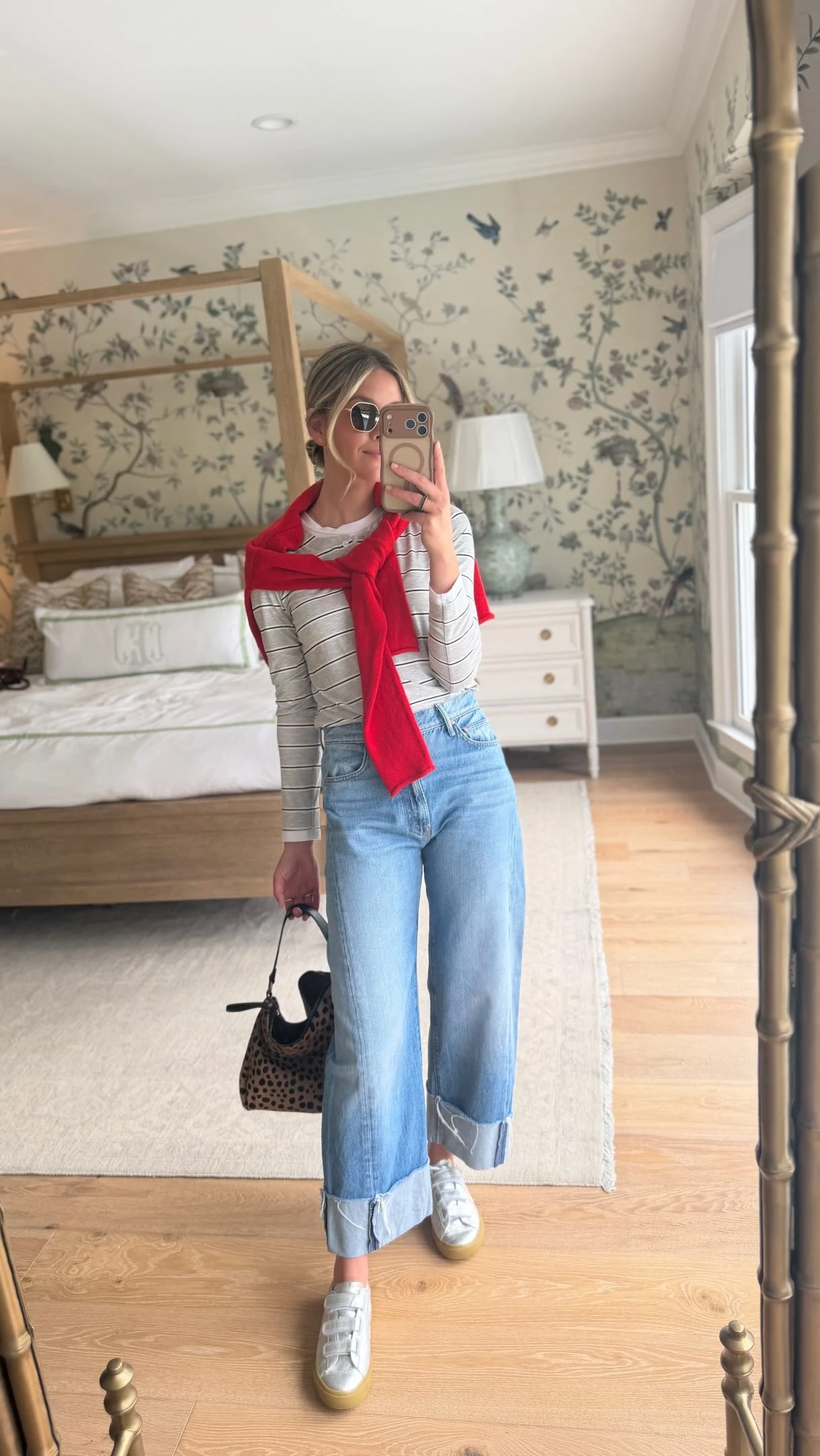 Red sweater tied over a striped shirt with wide-leg jeans, white sneakers, and a dark handbag.