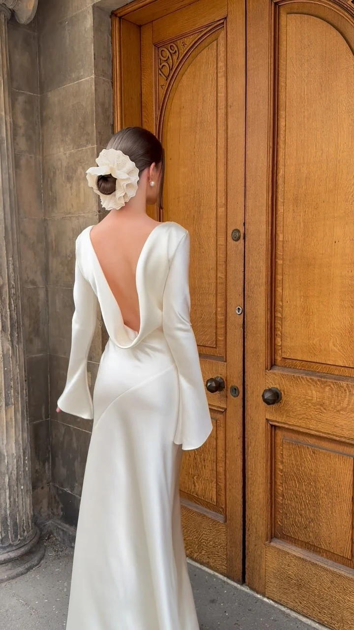 Floor-length white satin gown with boat neckline, bell sleeves, and dramatic open back.