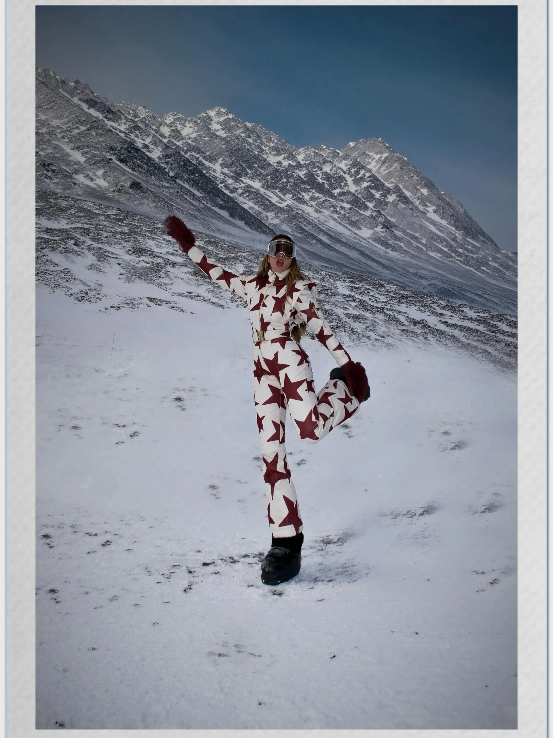 Person in a white ski jumpsuit with burgundy stars, belt, dark gloves, boots, and reflective goggles.