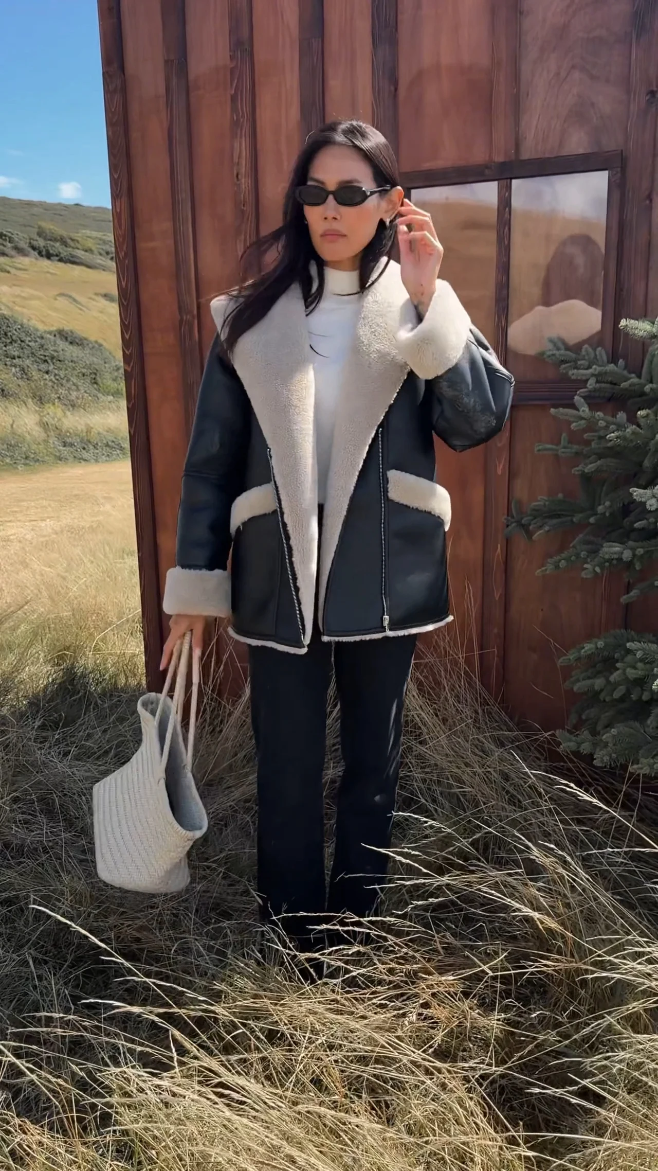 Black shearling jacket with cream trim worn over a white high-neck top, slim dark pants, and a woven tote.