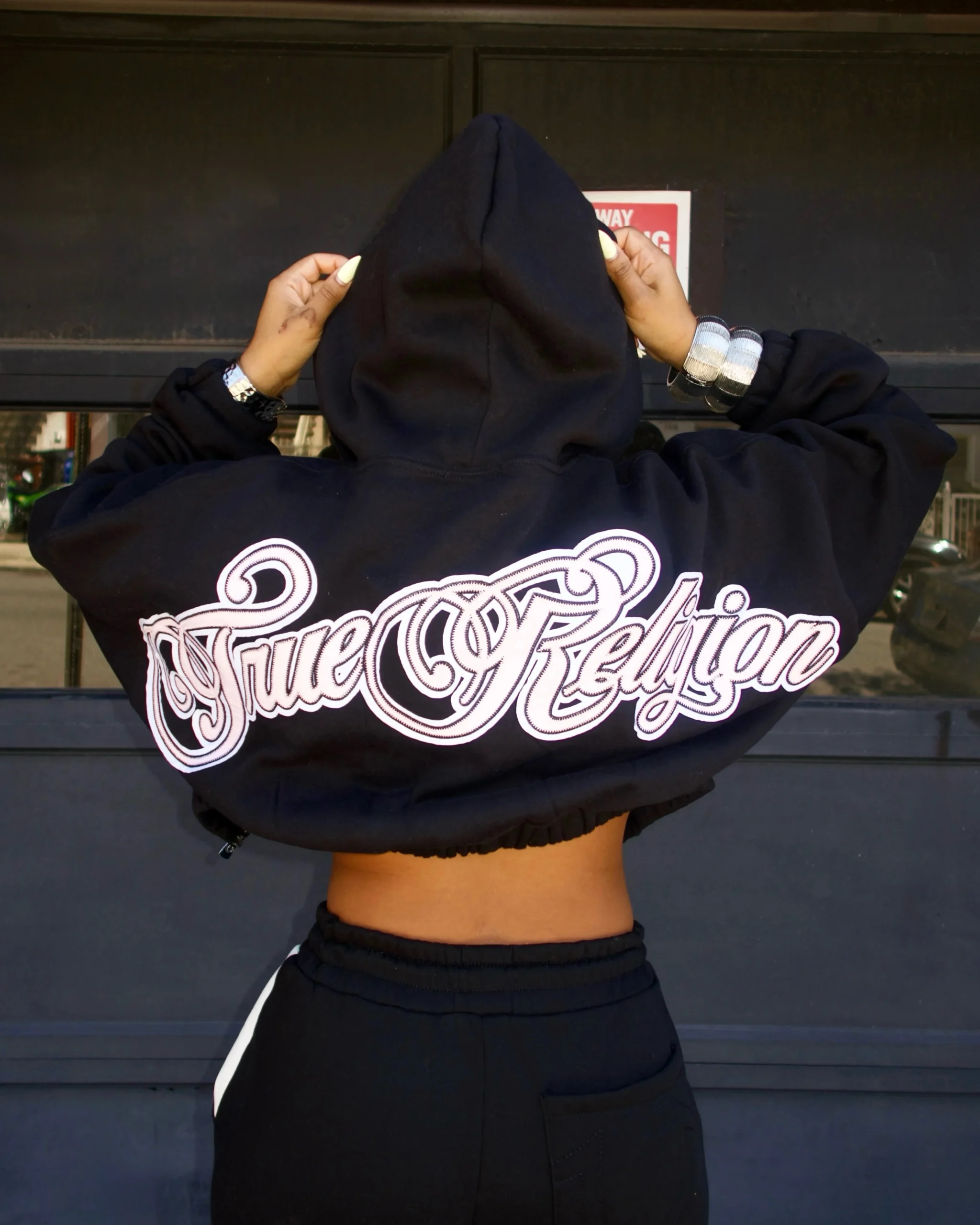 Person wearing a black cropped hoodie with white script and black track pants with white stripe.