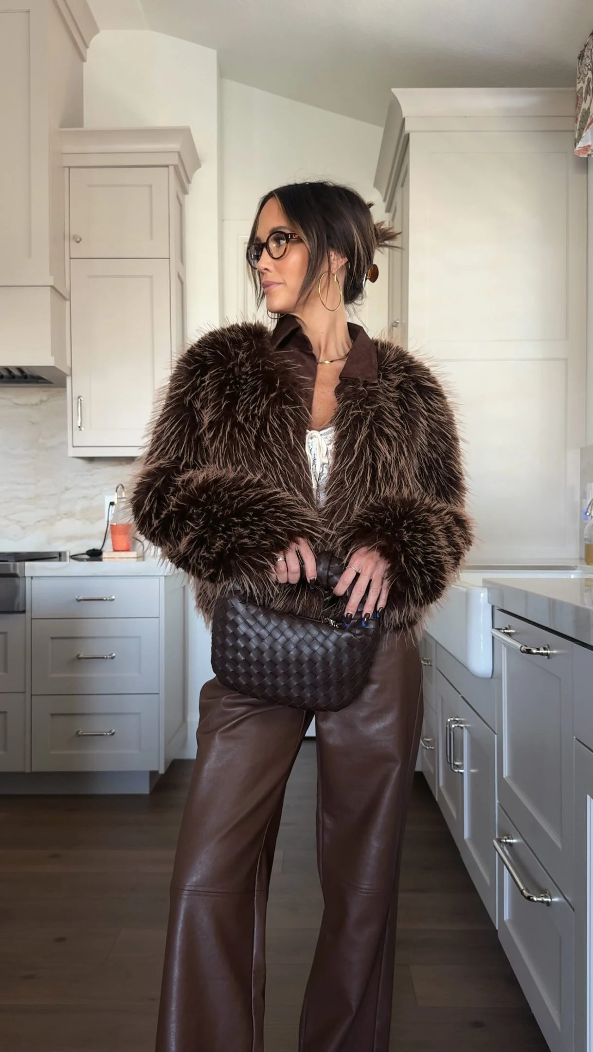 The outfit features a voluminous dark brown shaggy jacket with a broad collar, worn over a glimpse of a light-colored top. Smooth dark brown wide-leg pants fall to the floor. A dark woven handbag is held in front of the waist.