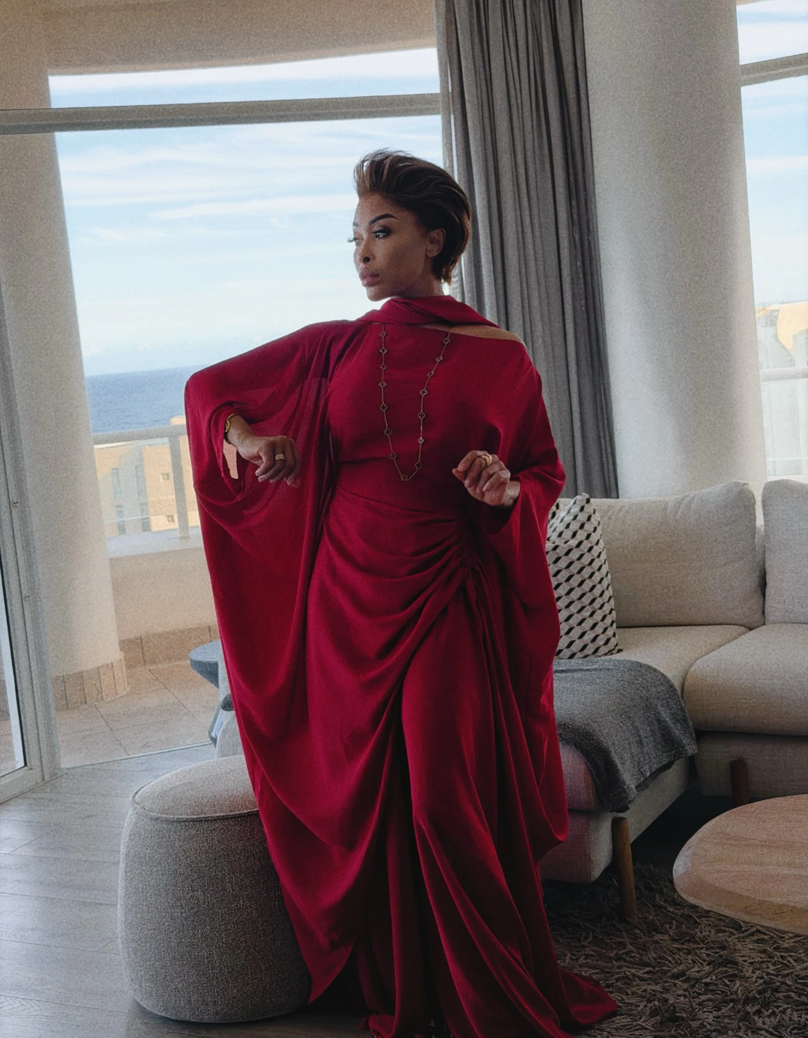 A floor-length cherry red dress has a high neckline, long cape-like sleeves and abundant draping gathered at the waist and hips. The fabric falls in loose folds over the legs, with a long slit visible near the front. A long, fine chain necklace hangs down the center of the dress. The outfit is shown without visible shoes, and no other clear accessories are apparent.