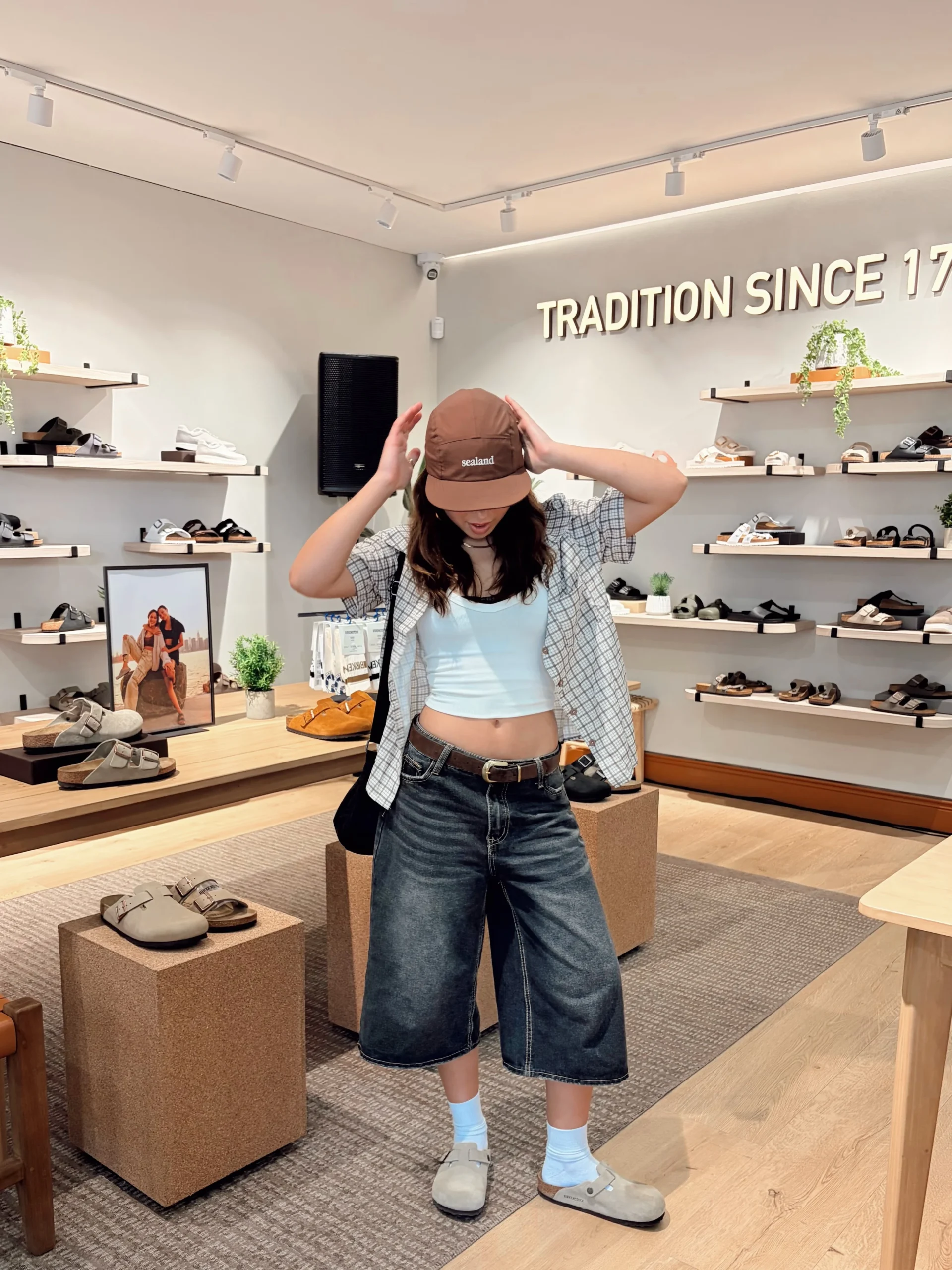 Person in a crop tank, open plaid shirt, baggy dark denim, white socks, and beige Boston clogs.
