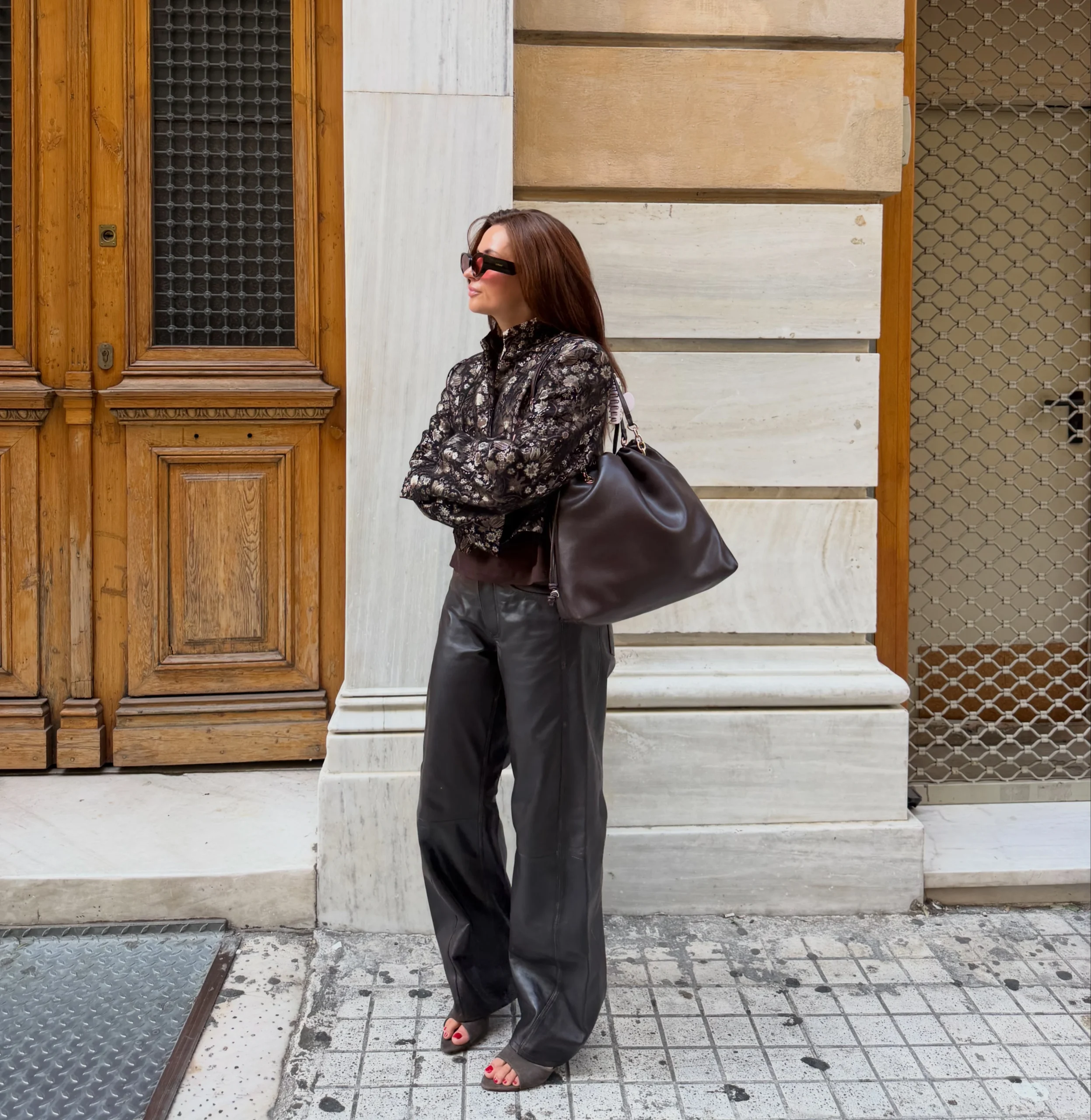 Dark brown short-sleeve T-shirt, black-and-gold floral cropped jacket, black wide-leg leather pants, large brown leather tote bag, dark sunglasses, heeled sandals