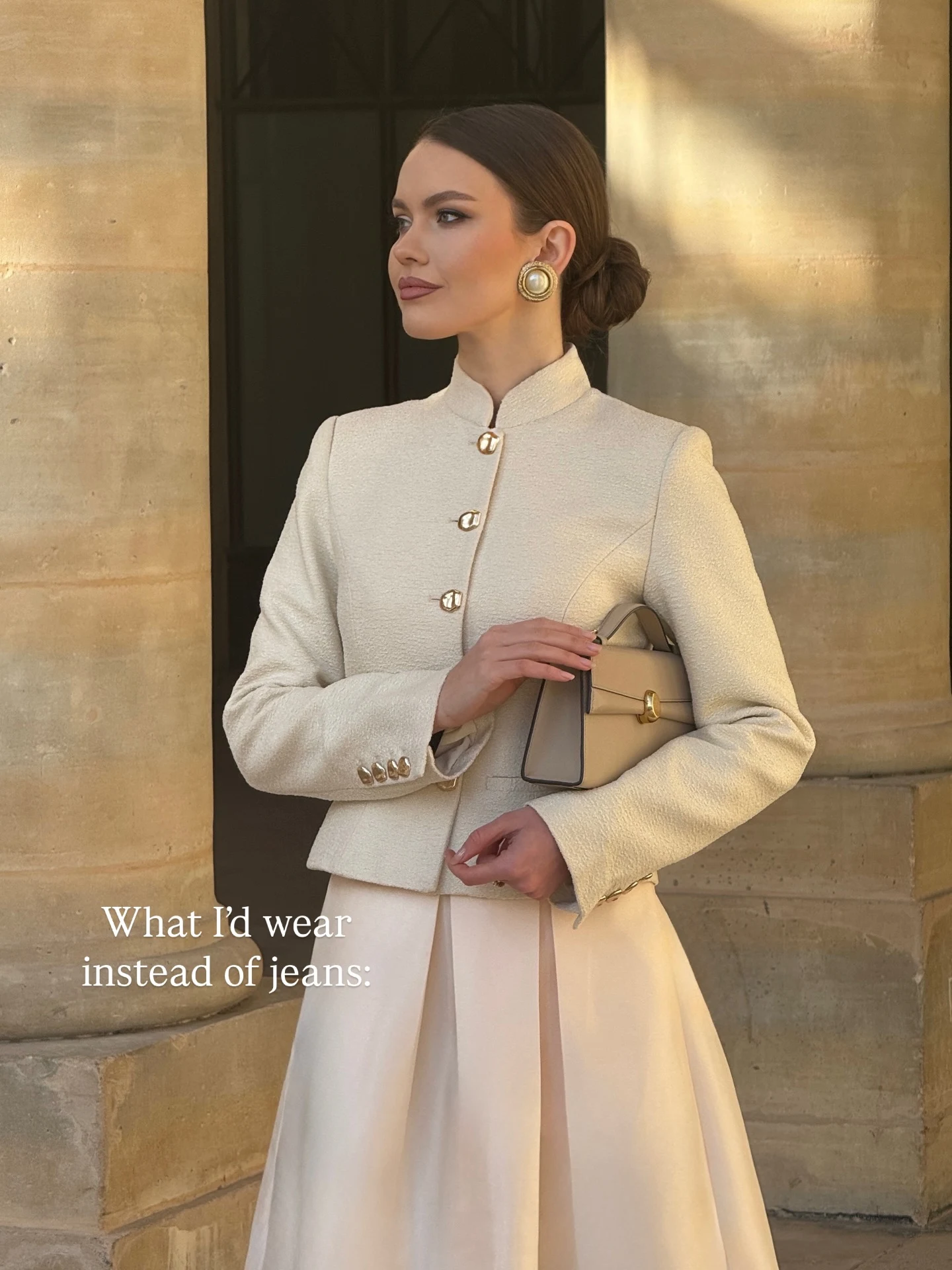 Cream collarless jacket and matching pleated skirt with a structured beige bag and gold earrings.