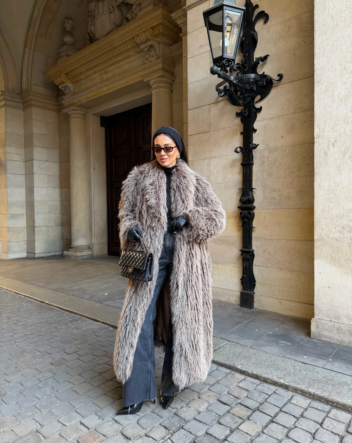 Woman in a long taupe shaggy faux-fur coat, black turtleneck, wide-leg jeans, and black gloves.