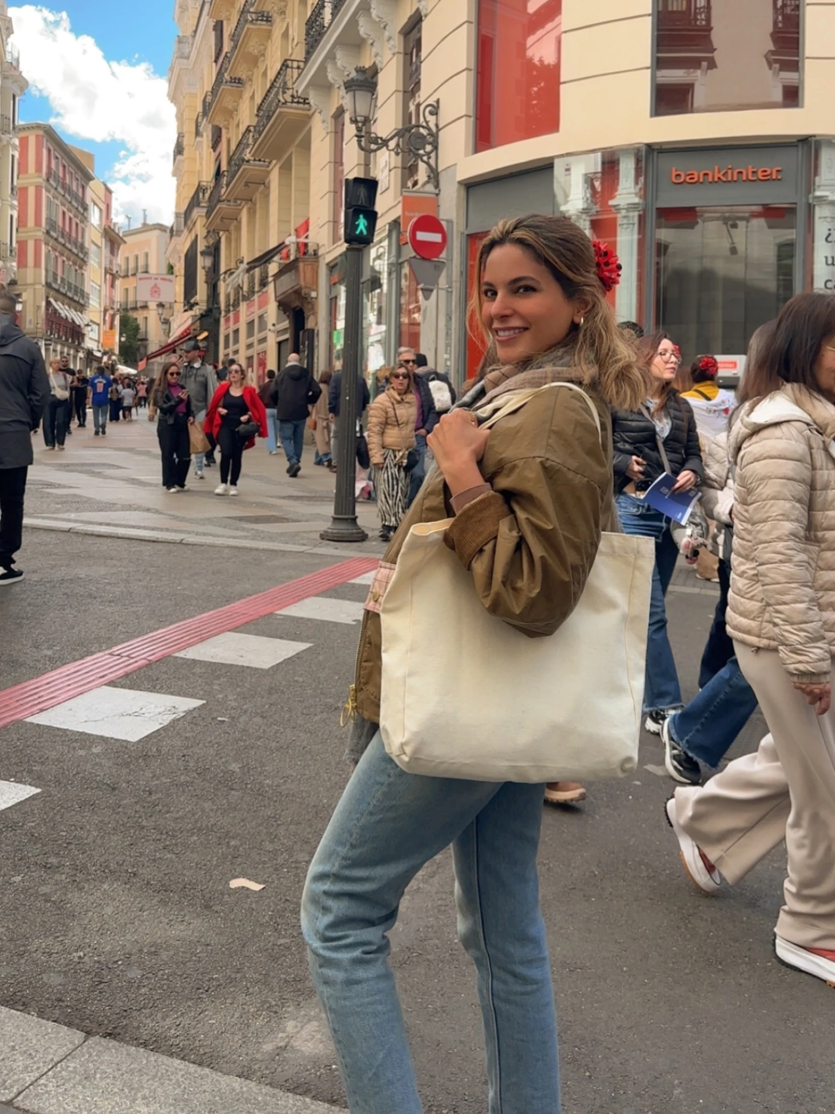Olive jacket, pale blue straight-leg jeans, cream tote, and sneakers styled on a busy street.