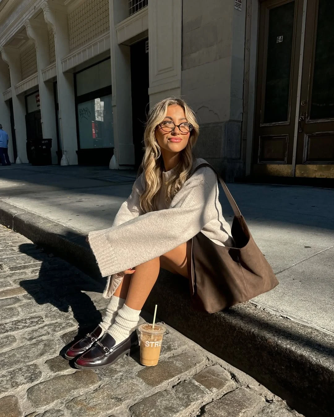 Woman in an oversized beige knit sweater, dark mini skirt, white socks, loafers, and brown tote.