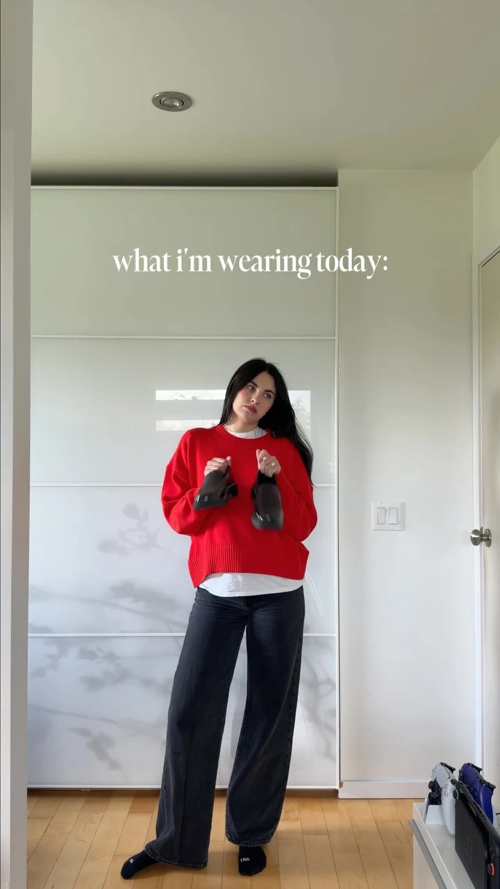 Red ribbed sweater over a white shirt with dark wide-leg trousers, black socks, and dark shoes held.