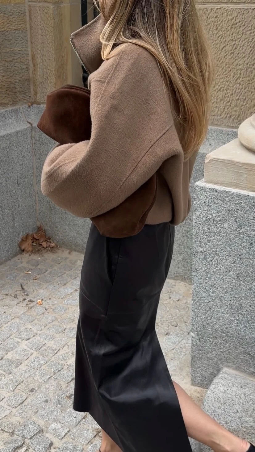 Person in a long taupe coat, black skirt, brown clutch, and black open-toe shoes.
