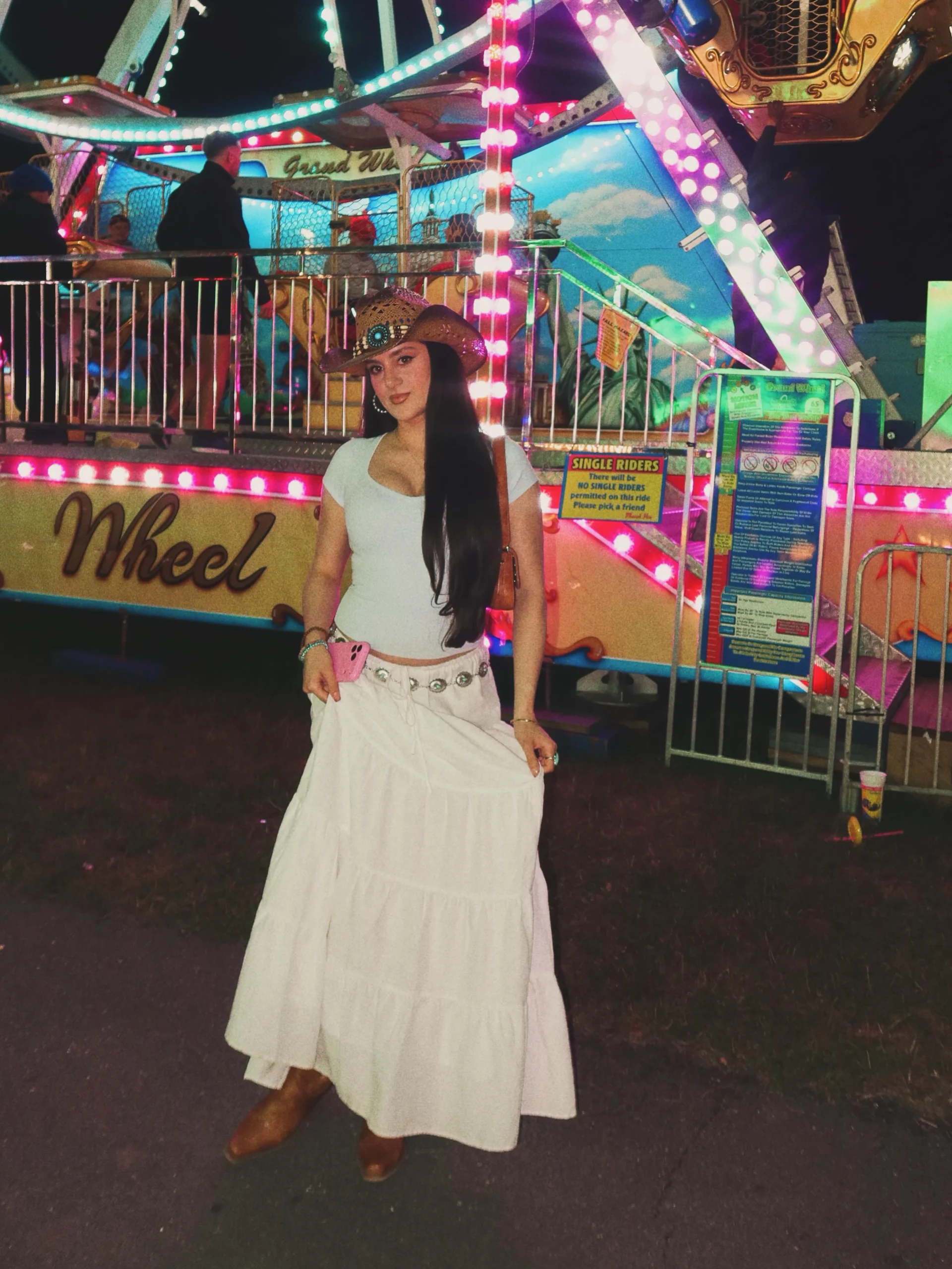 Woman in a white crop top, tiered maxi skirt, concho belt, cowboy hat, and brown boots.