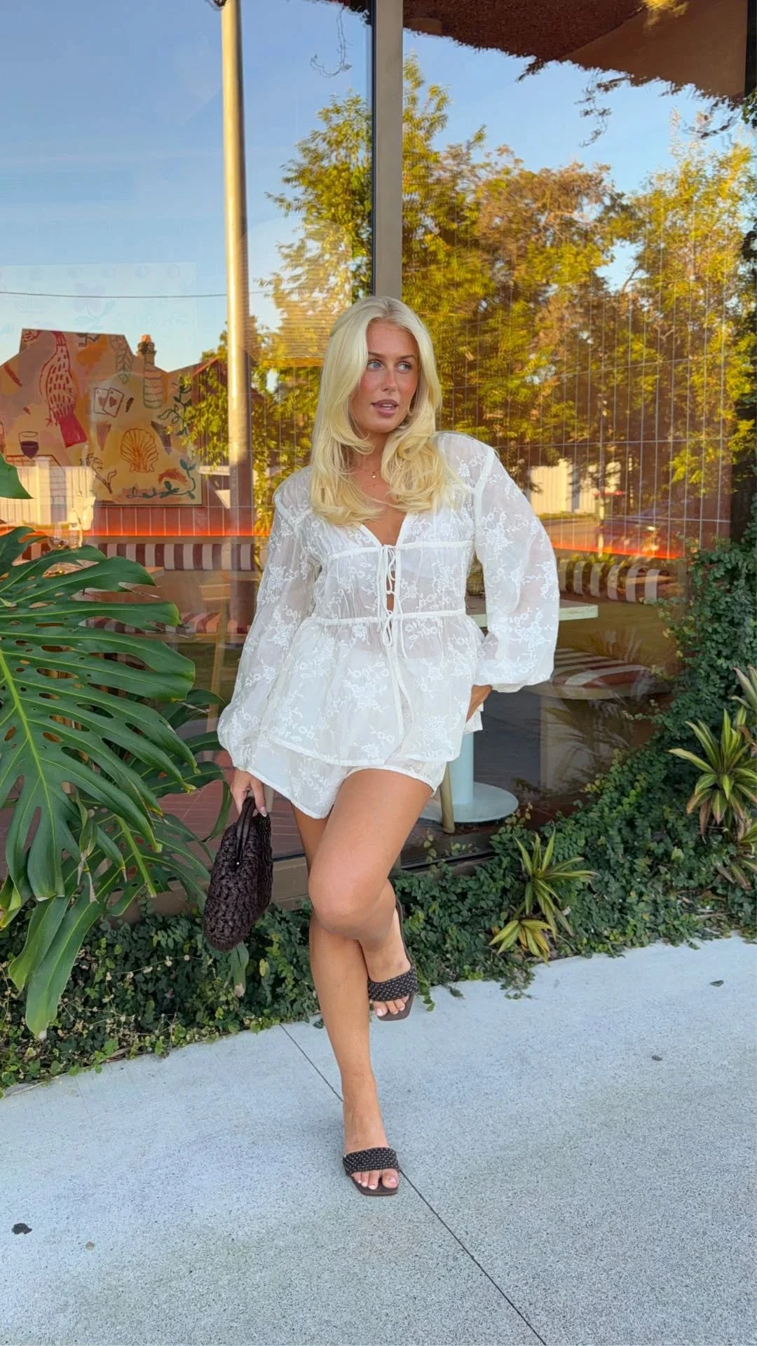 The outfit consists of a white blouse with a floral lace pattern, a V-shaped neckline, front ties, and long loose sleeves, paired with matching white shorts. The blouse has a gathered waist and a slightly flared hem. Black open-toe sandals and a dark woven handbag are also visible.