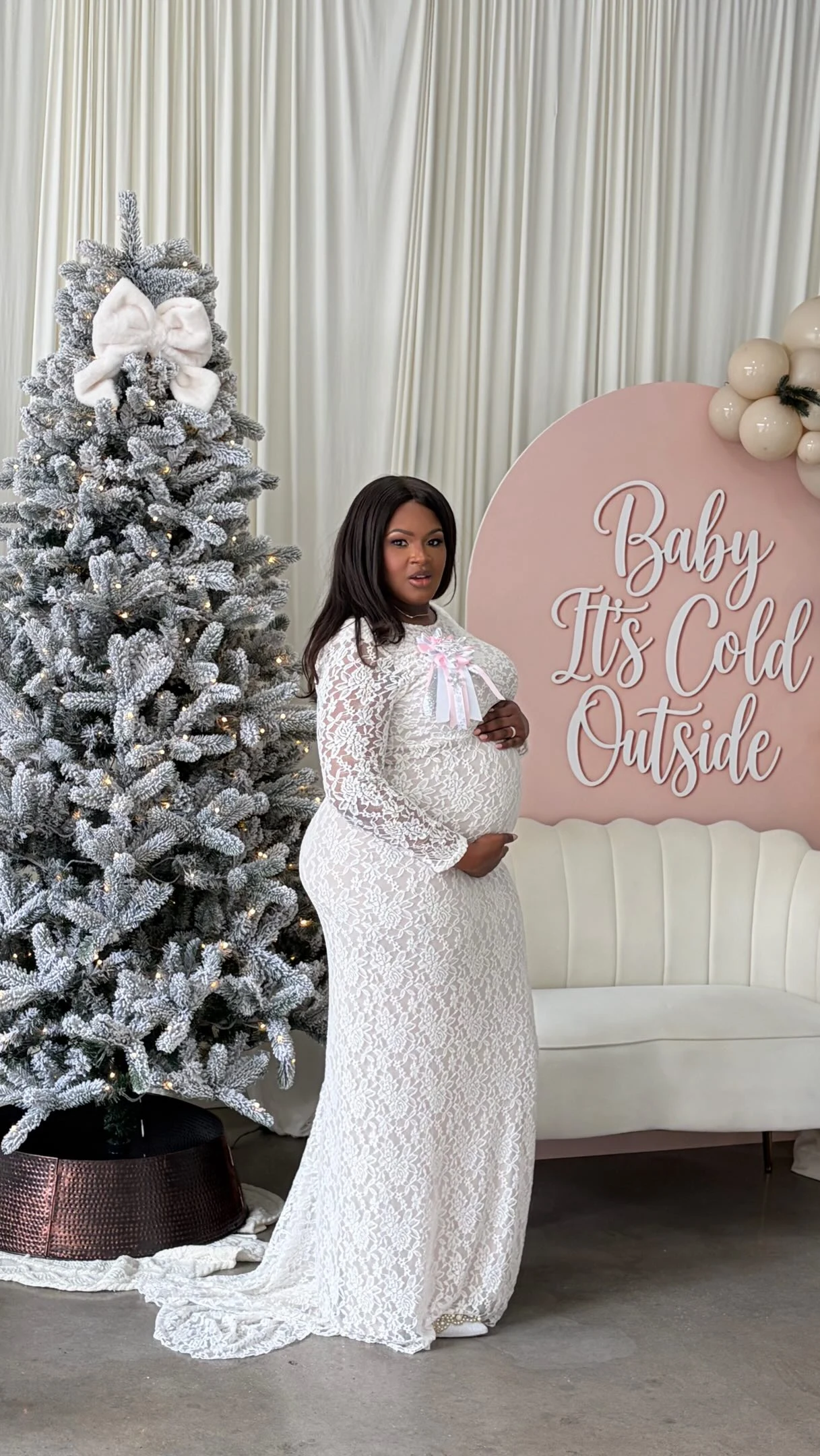 Pregnant woman in a fitted white lace gown with long sleeves and a pale pink ribbon bow at the neckline.