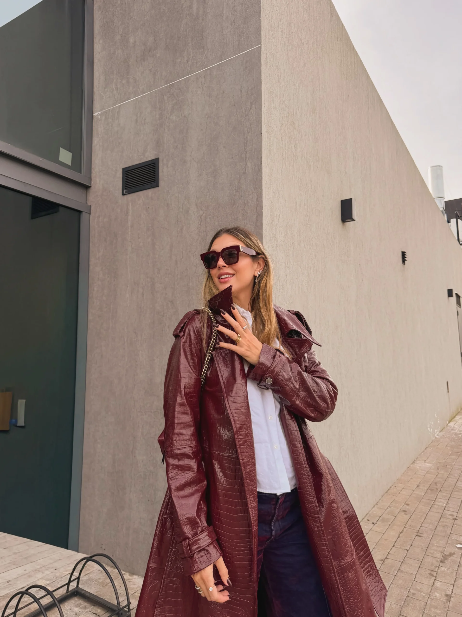 The outfit features a long burgundy coat with a glossy finish, wide lapels, and a matching belt tied at the waist. A plain white top shows beneath it, paired with dark blue jeans. A narrow chain strap crosses one shoulder, and the person wears oversized dark sunglasses. The coat falls below the hips and is worn open over the top and jeans. Shoes are outside the frame.
