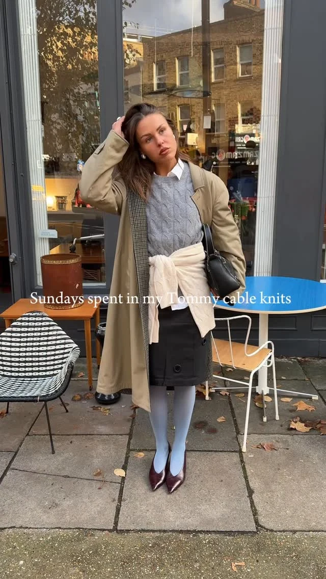 Woman wearing a cable-knit sweater, black mini skirt, light blue tights, burgundy heels, and a beige trench coat.