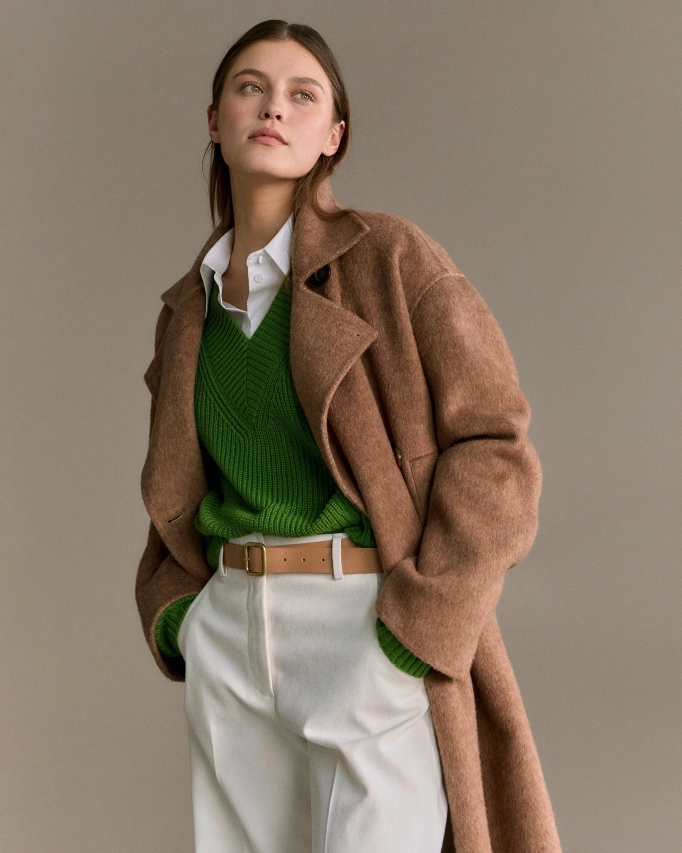 Person in a camel coat, green V-neck sweater, white shirt, and high-waisted white pleated trousers.
