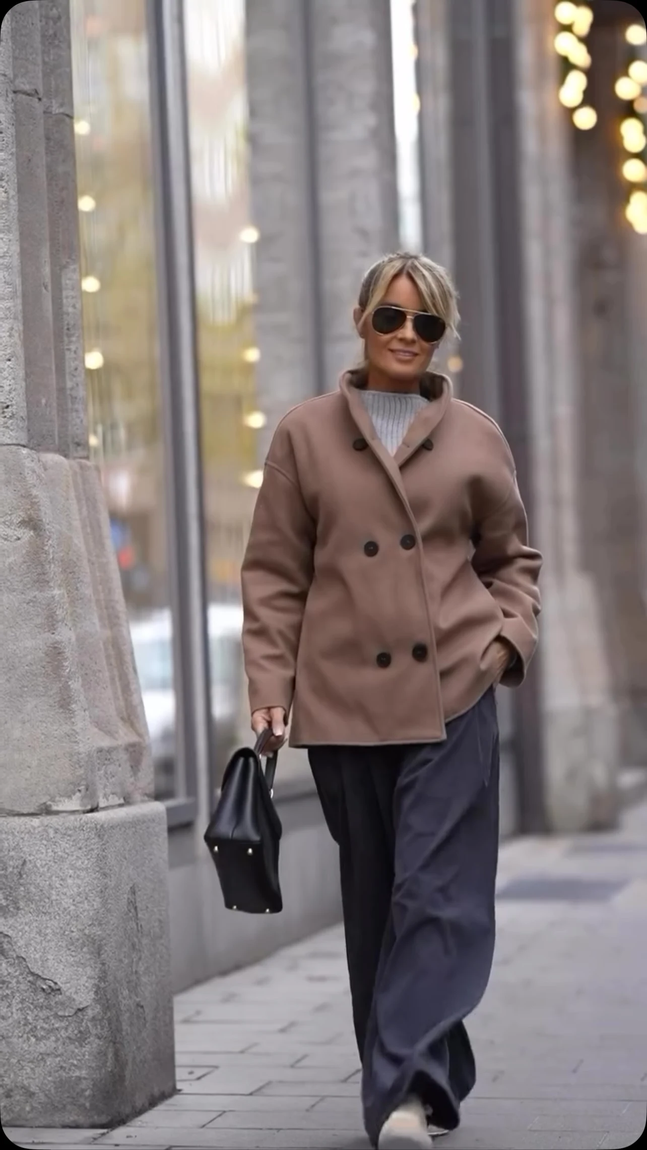 Camel double-breasted wool coat over light gray ribbed sweater, dark wide-leg trousers, small black handbag