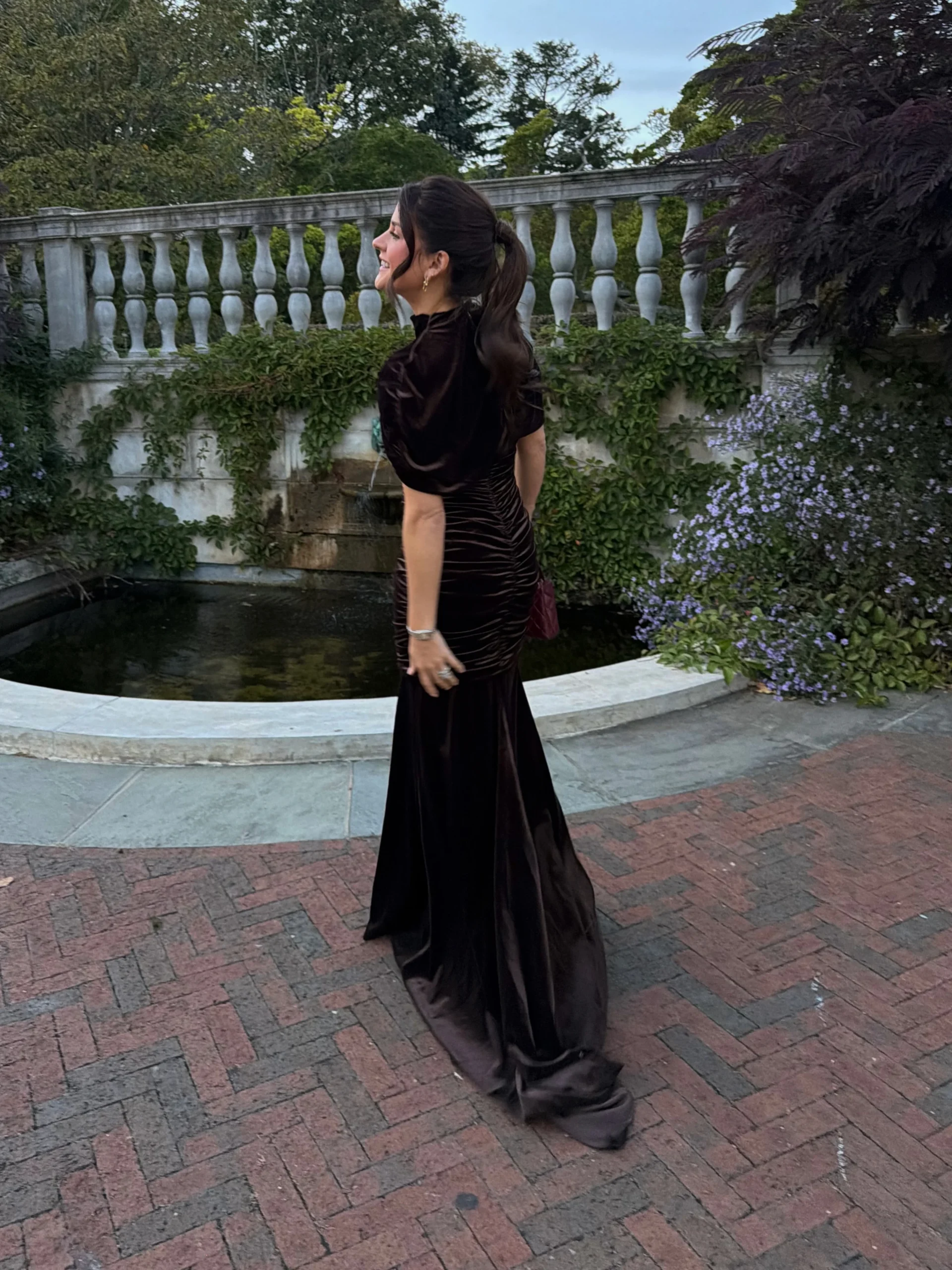 Dark nearly black ruched gown with high neckline, short sleeves, and a trailing flared skirt beside garden greenery.