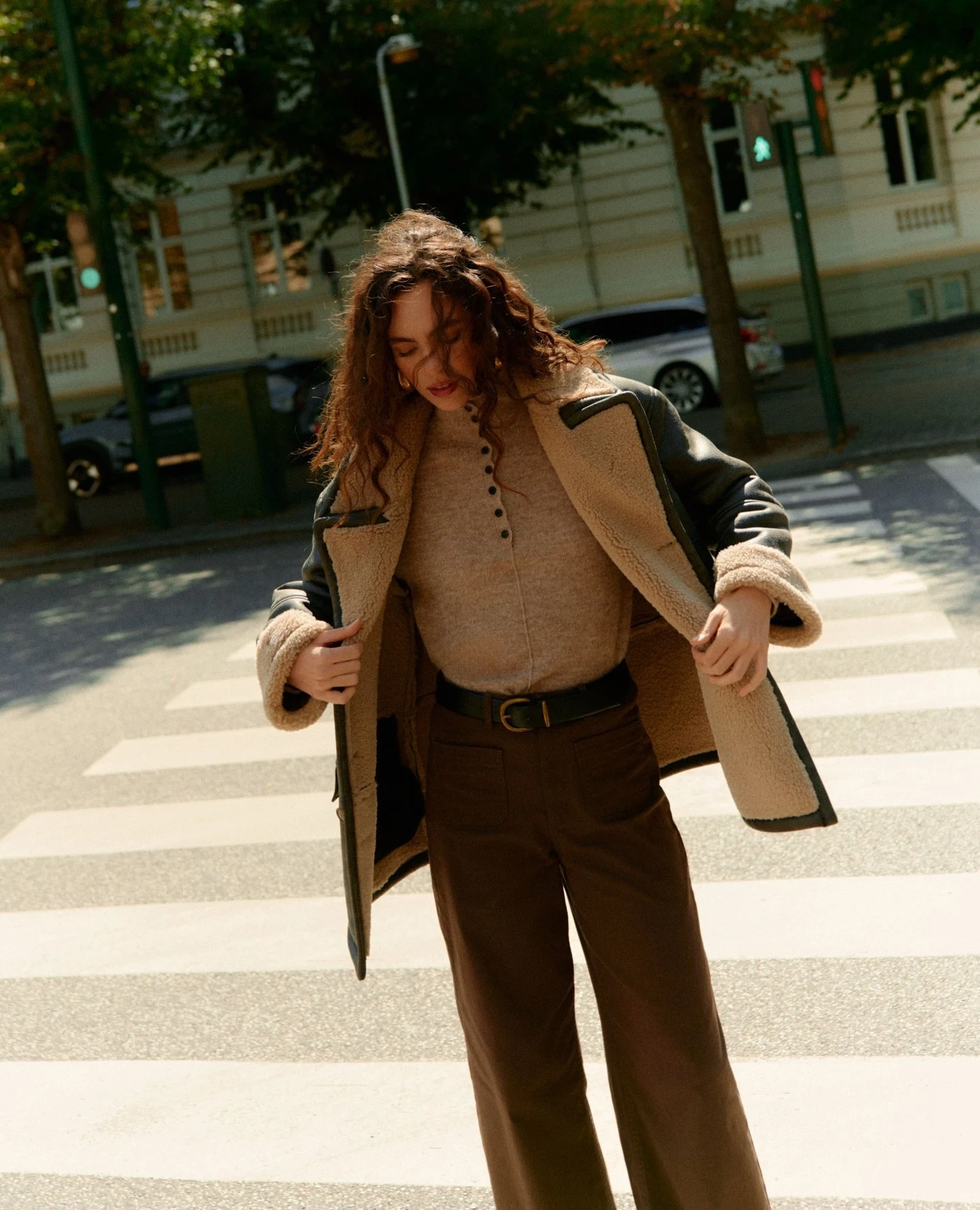 Long dark shearling jacket with tan lining worn over a beige top, wide-leg brown trousers, and a black belt.