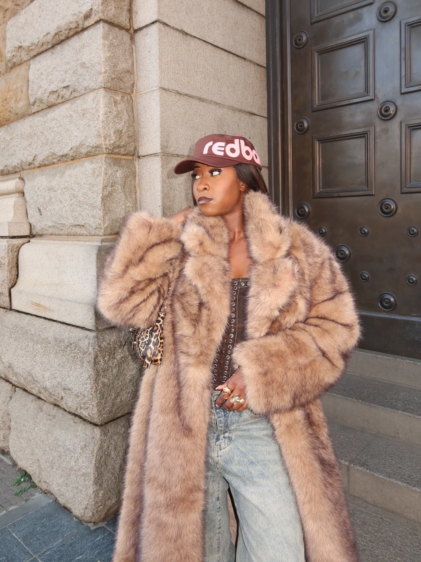 Woman wearing a light-brown faux-fur coat, corset top, baggy jeans, heeled boots, and a leopard crossbody bag.