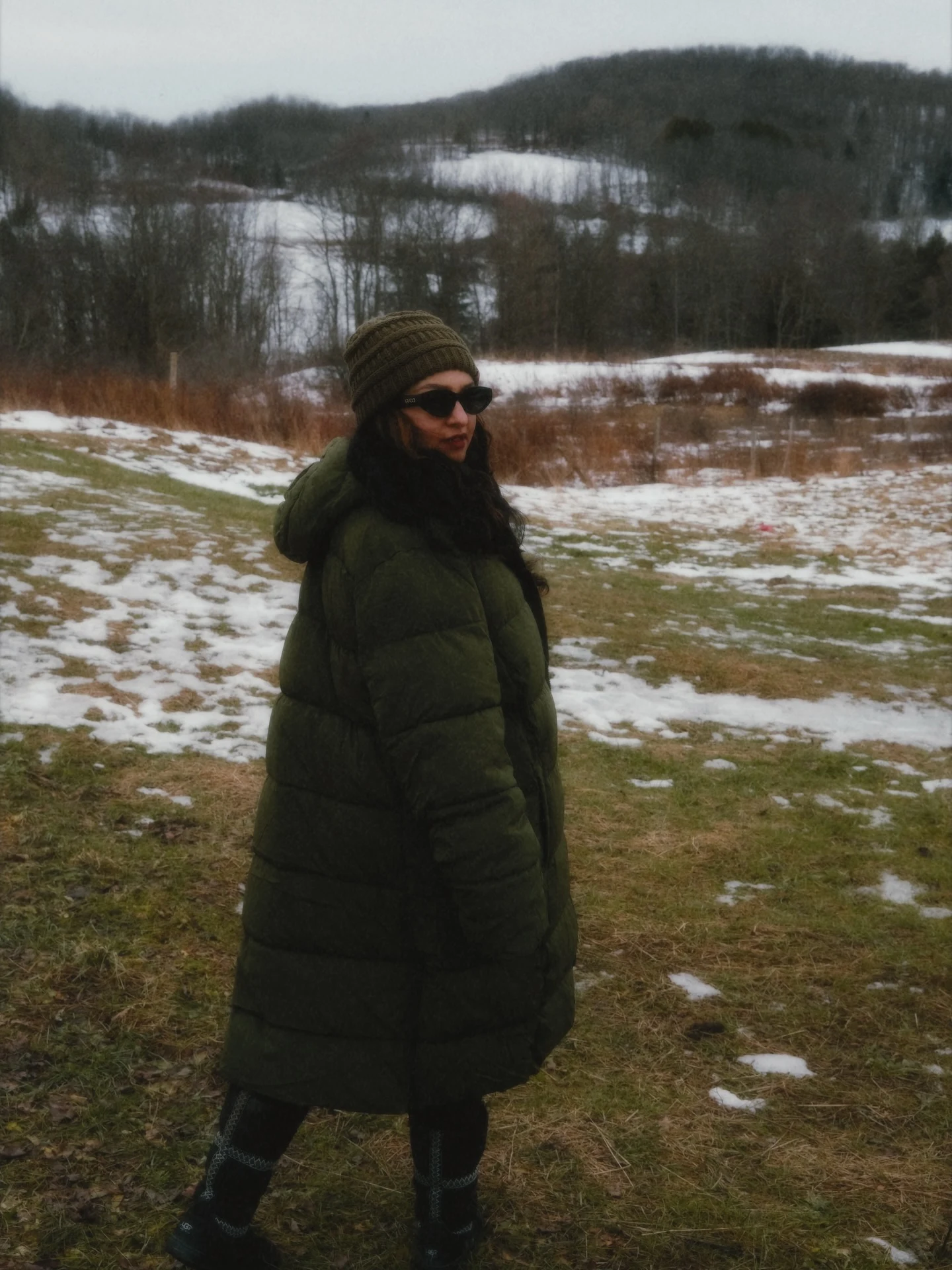Long olive hooded puffer coat worn with a ribbed knit beanie and dark sunglasses outdoors in snow.