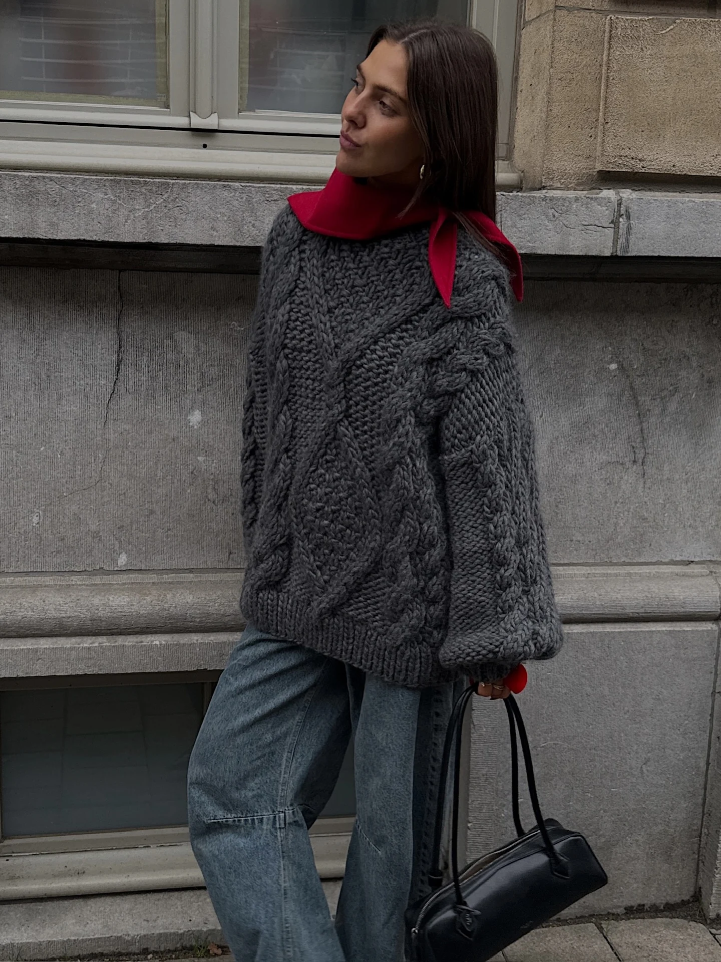 Dark gray cable-knit oversized sweater with a red scarf, blue-gray jeans, and a black handbag.