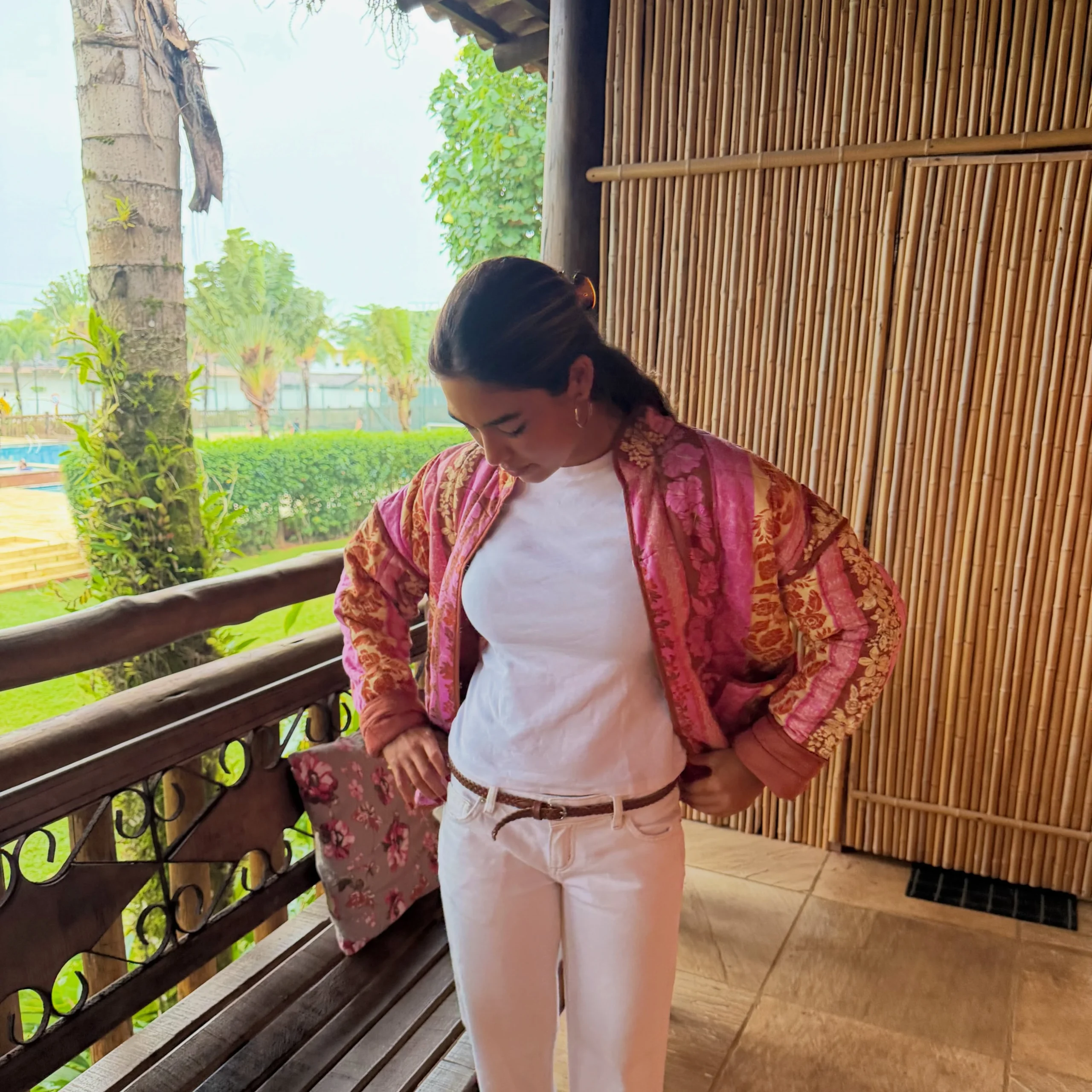 Person wearing an open pink patterned jacket over a white top and white trousers with a narrow brown belt.