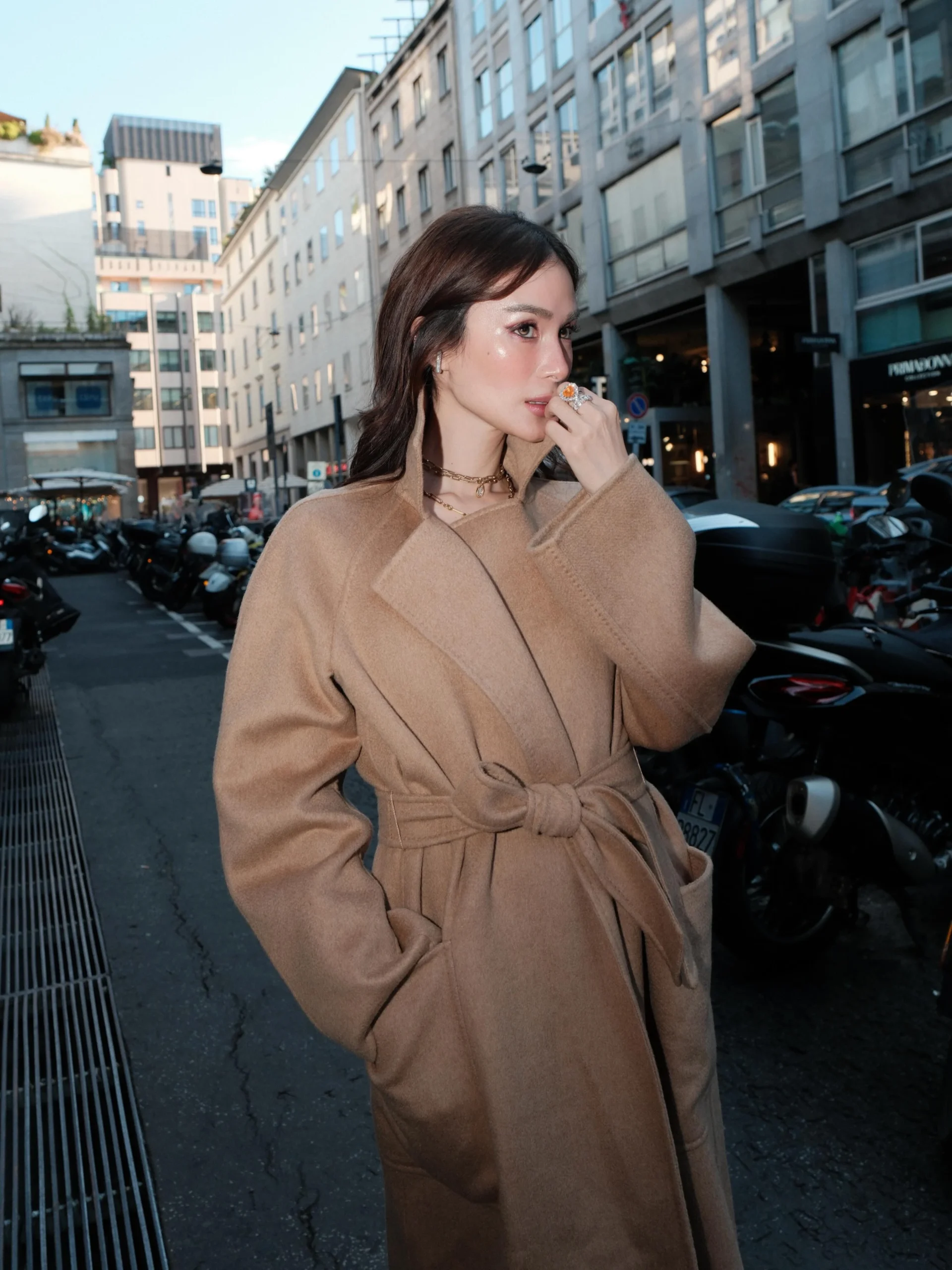 Long belted camel wool coat styled with tailored trousers and layered gold necklaces.