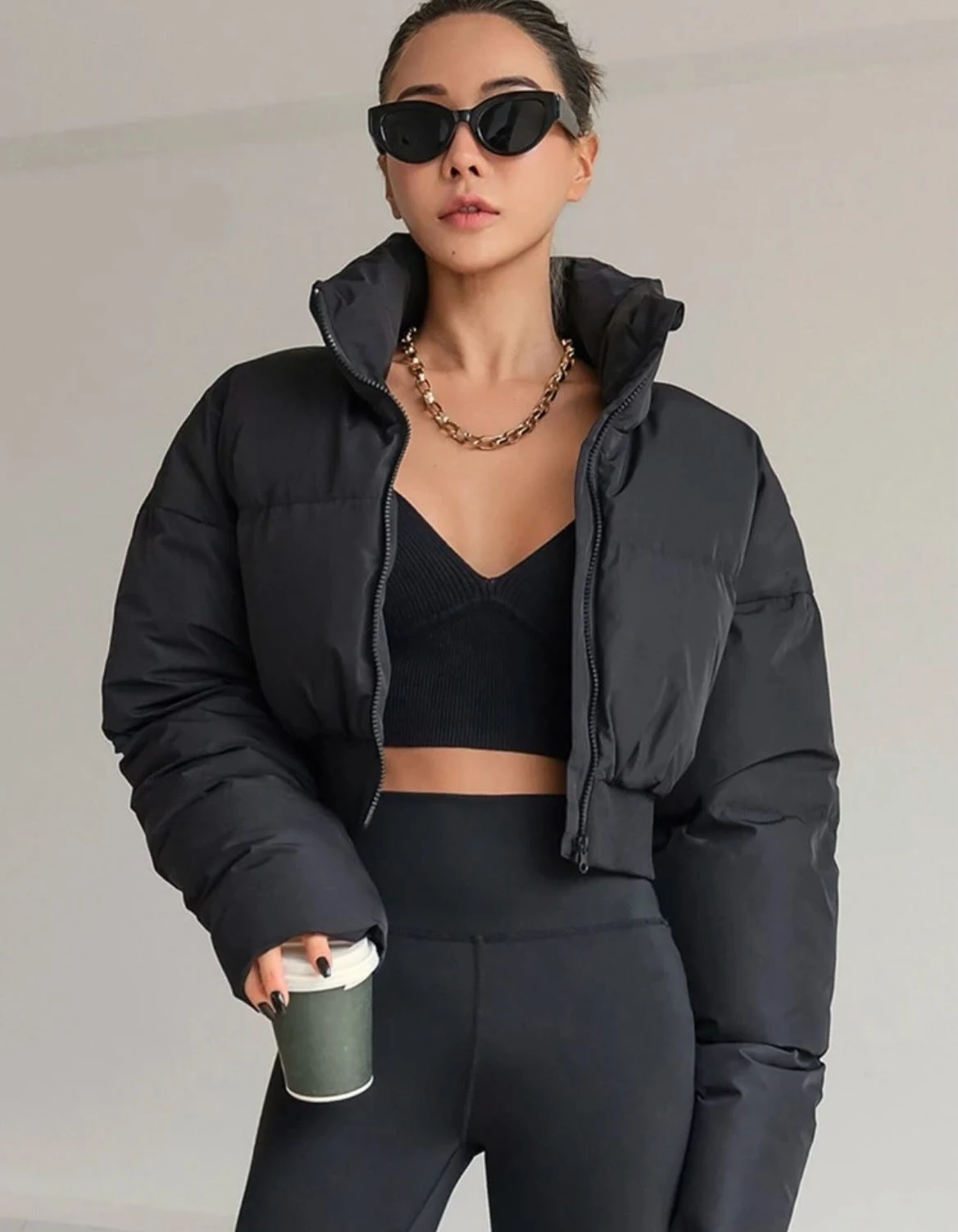Black cropped puffer jacket worn open over a ribbed crop top, black leggings, gold chain, and angular sunglasses.