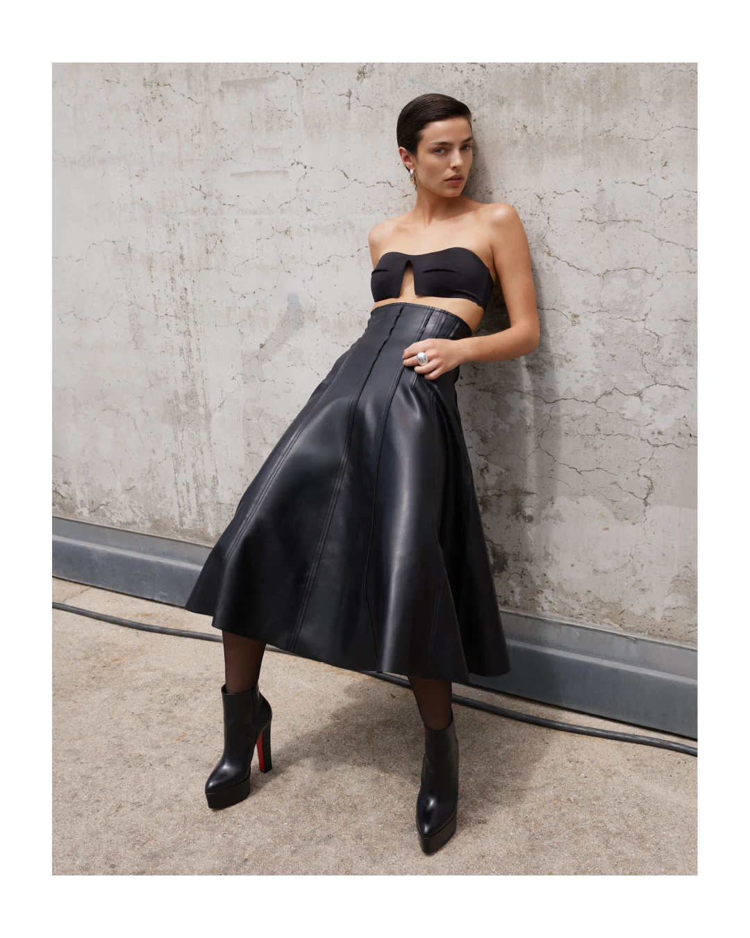 Black leather midi skirt, bandeau top, sheer tights, and platform ankle boots in an all-black outfit.