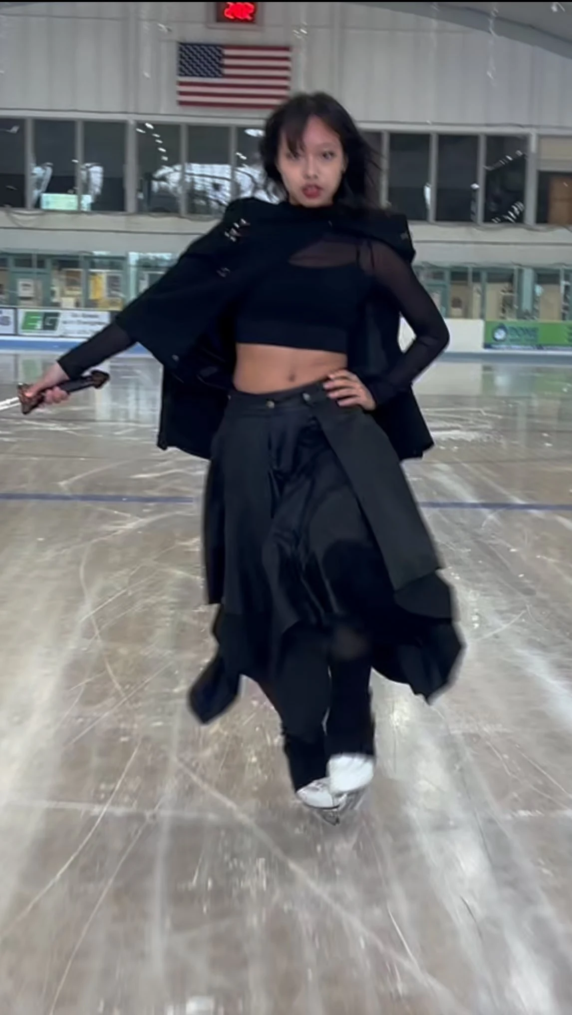 Skater in a black sheer crop top, cape layer, layered skirt, and white ice skates on the rink.