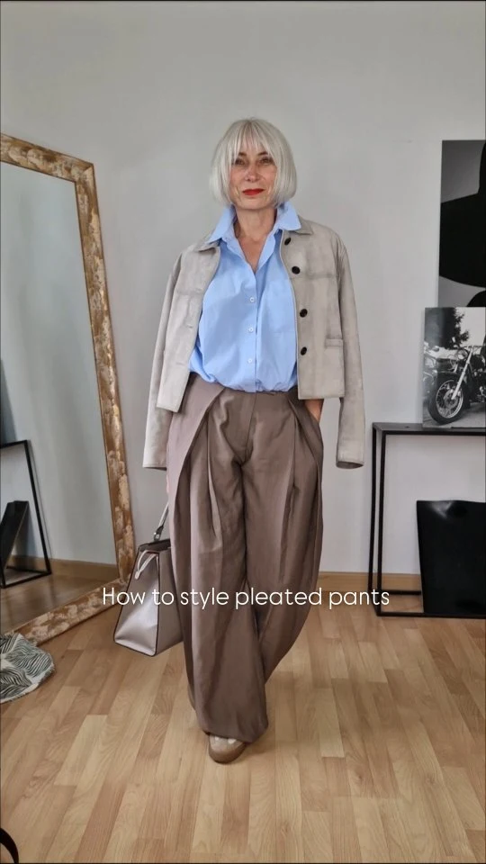 Blue button shirt and taupe pleated trousers under an open beige jacket with beige shoes and a light bag.