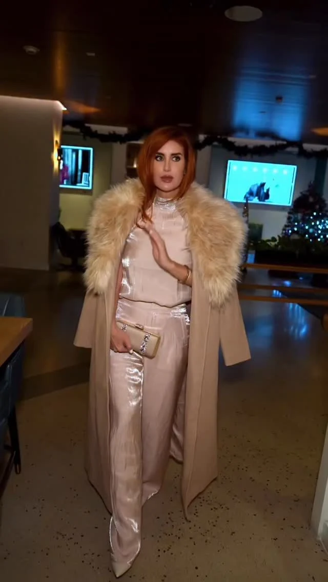 Woman wearing a blush satin wide-leg jumpsuit, long camel coat with fur collar, and cream crystal clutch.