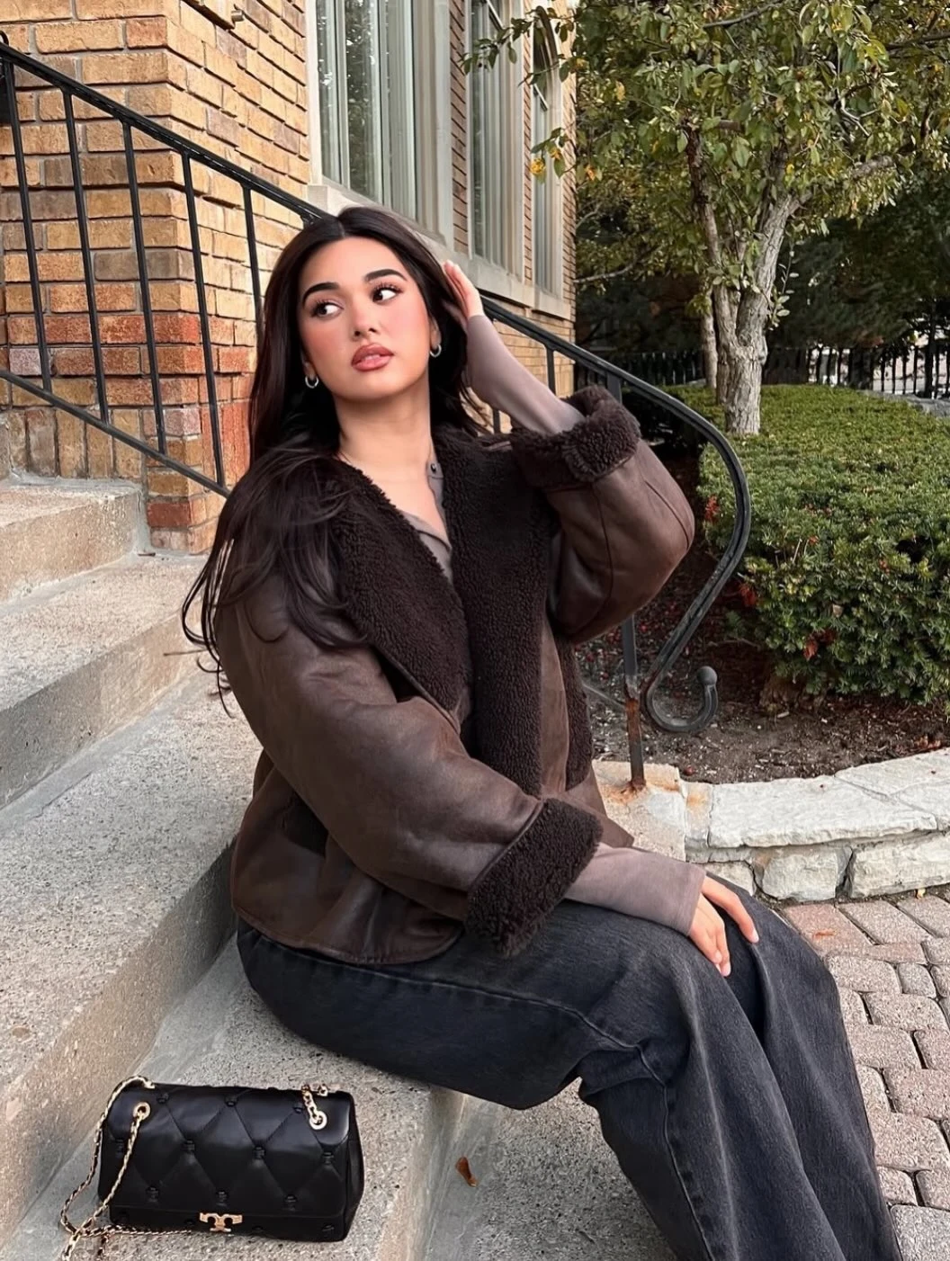 Dark brown shearling jacket with textured lapels worn over dark gray wide-leg jeans, with a black quilted bag.