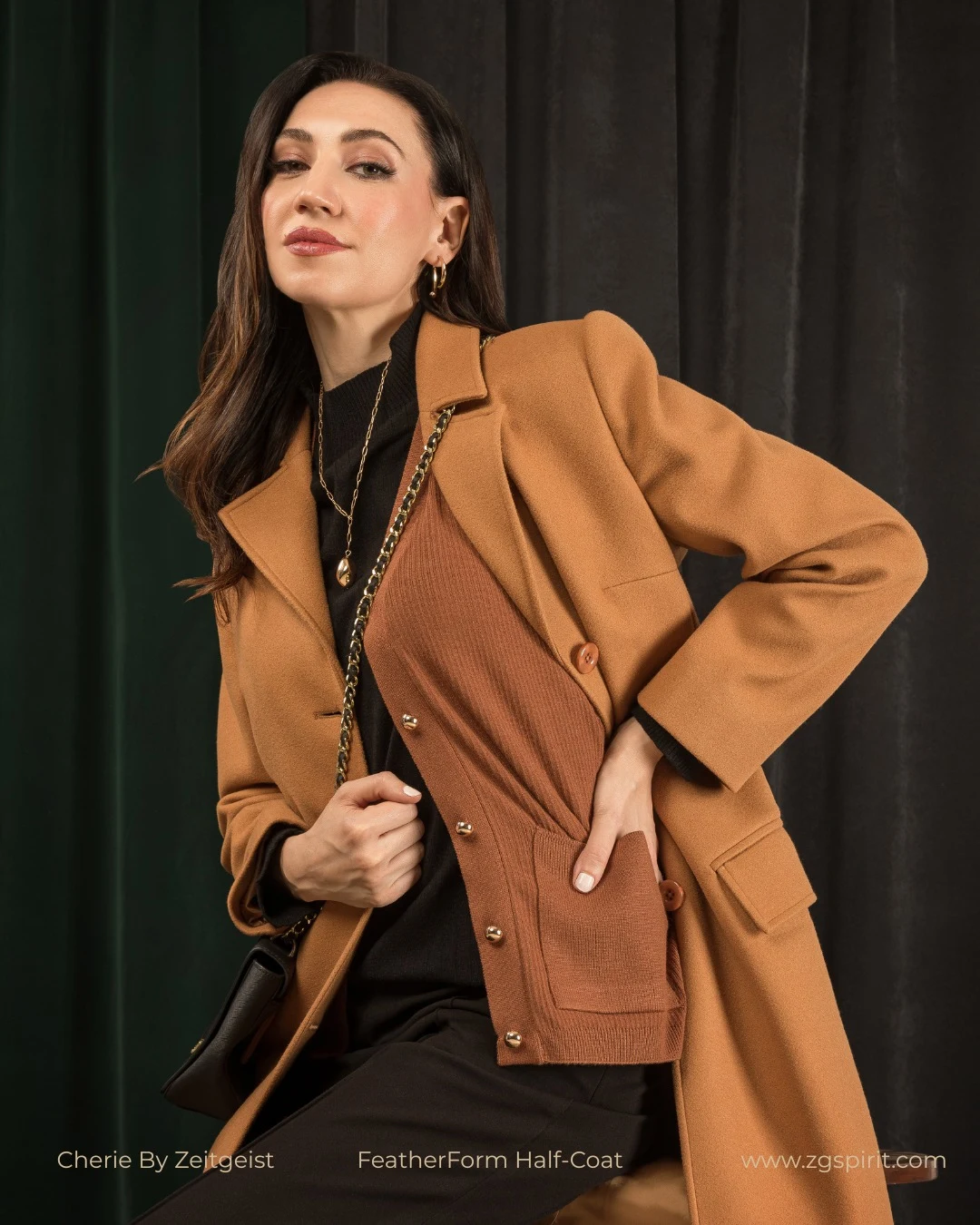 Person in a camel coat, rust-brown cardigan, black top, trousers, and a chain-strap shoulder bag.