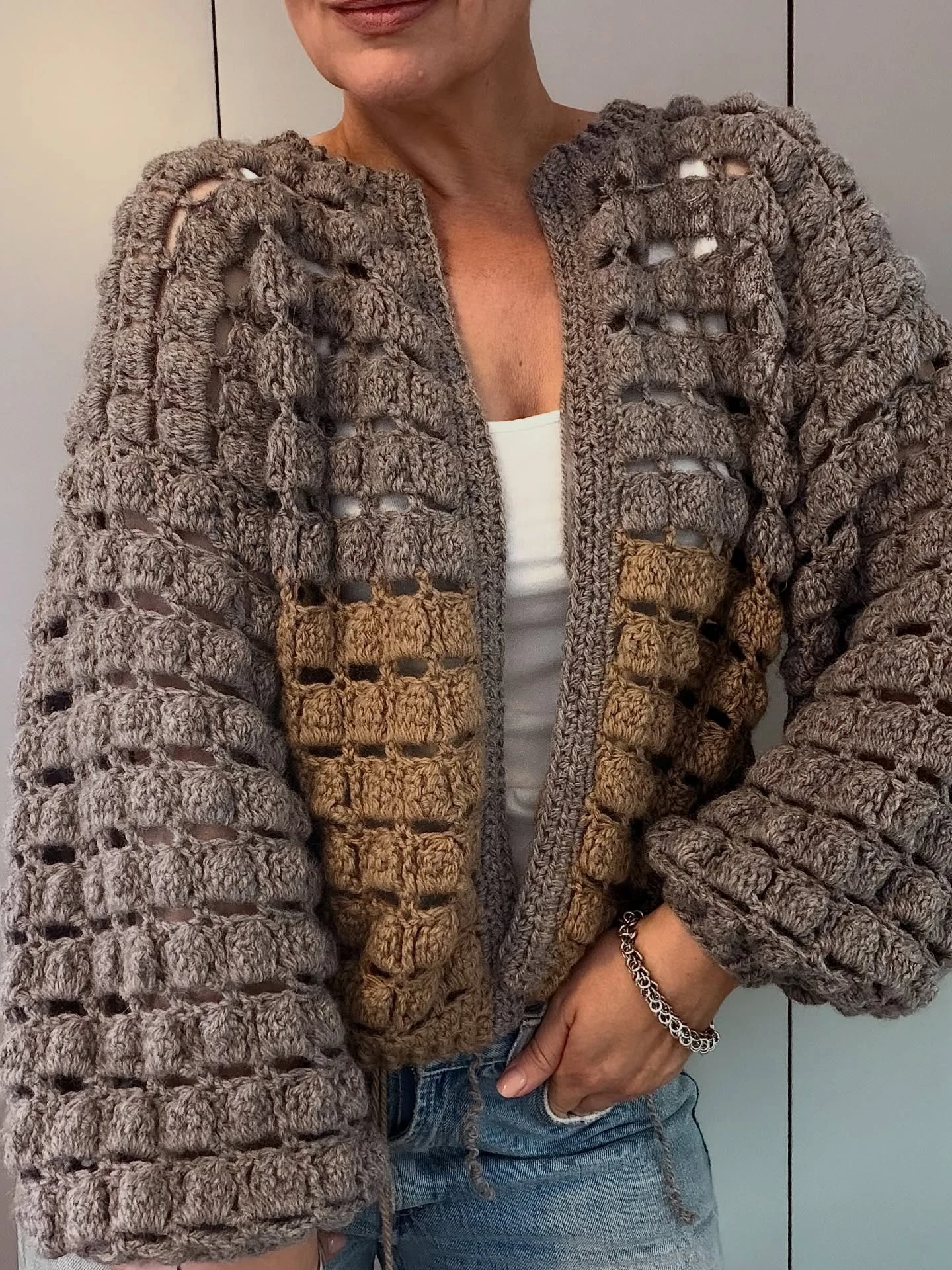 Cropped gray and tan crochet cardigan with openwork stitches over a white top and blue jeans.