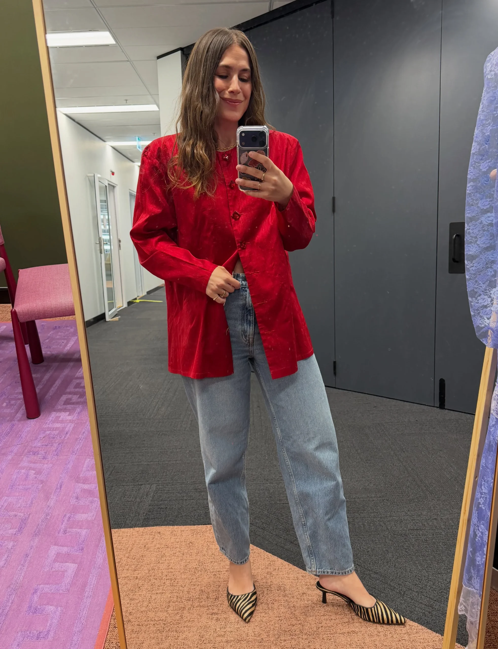 A bright red, long-sleeved button-front jacket is worn open over a dark top. The jacket has a loose, hip-length shape and a notched collar. Light blue jeans have a relaxed, wide-leg cut and reach the ankles. Pointed flats with narrow black and tan stripes complete the outfit. A thin necklace is visible at the neckline. The person holds a phone in front of the upper body, partially obscuring the top.