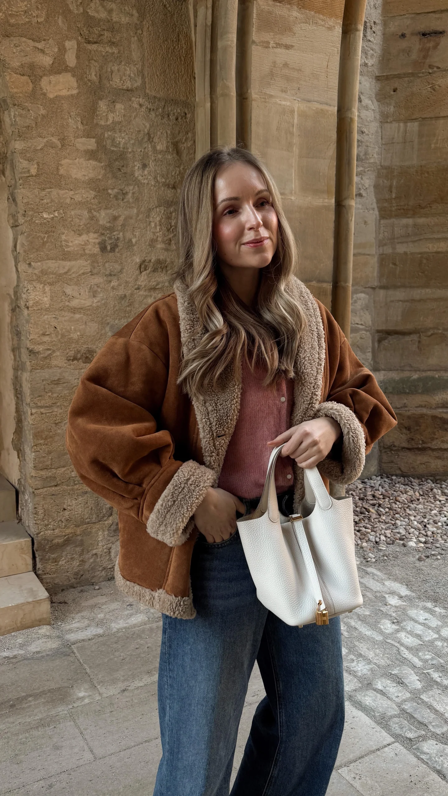Brown shearling-lined jacket with fur trim, pink knit button-up shirt, dark blue wide-leg denim jeans, brown pointed-toe ankle boots, white triangular tote bag