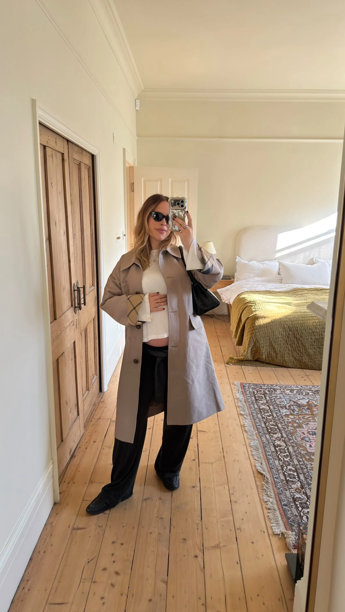 Person wearing a long taupe trench coat open over a white top with relaxed wide-leg black trousers.
