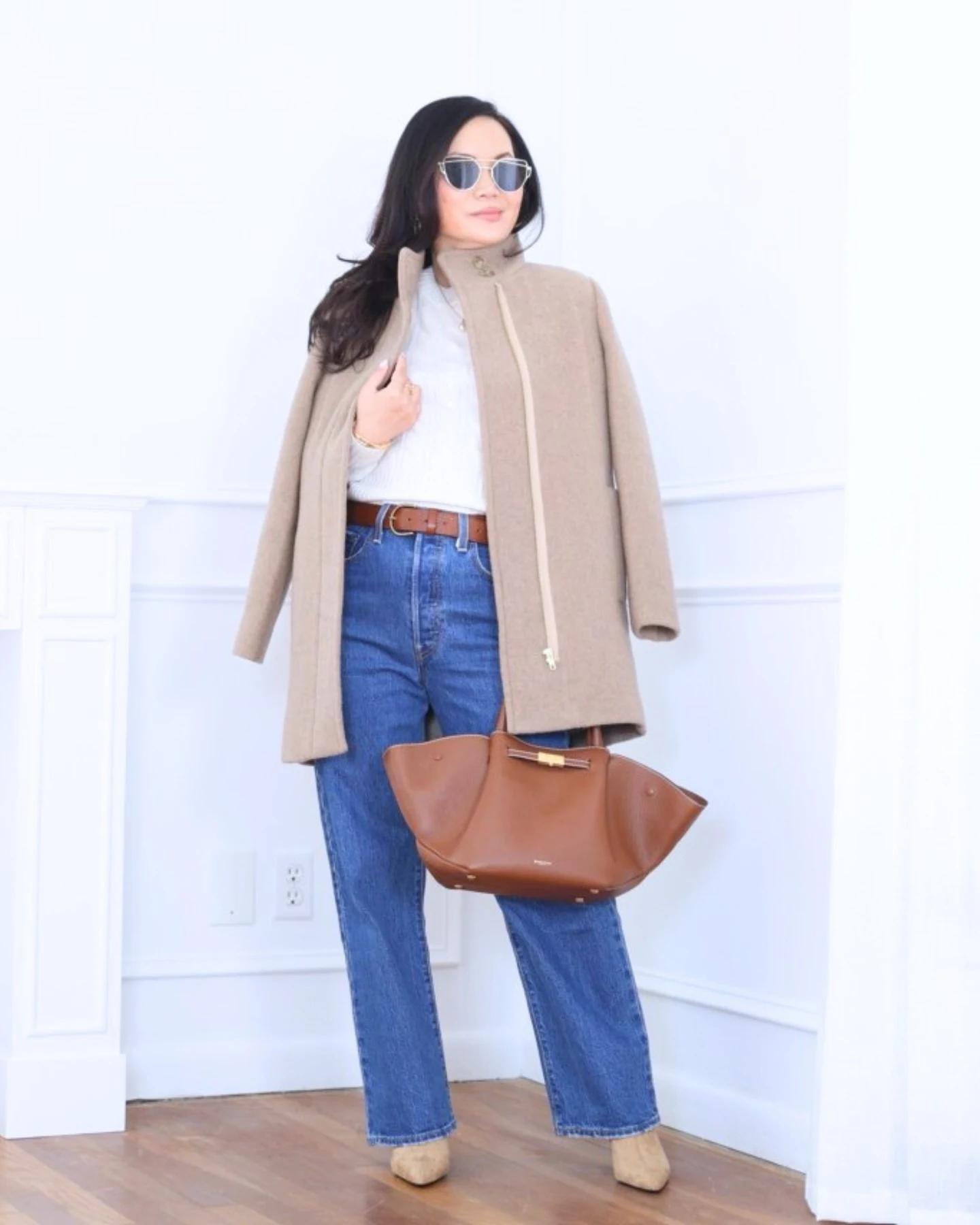 Petite woman wearing a beige city coat, white ribbed sweater, blue jeans, and tan ankle booties.
