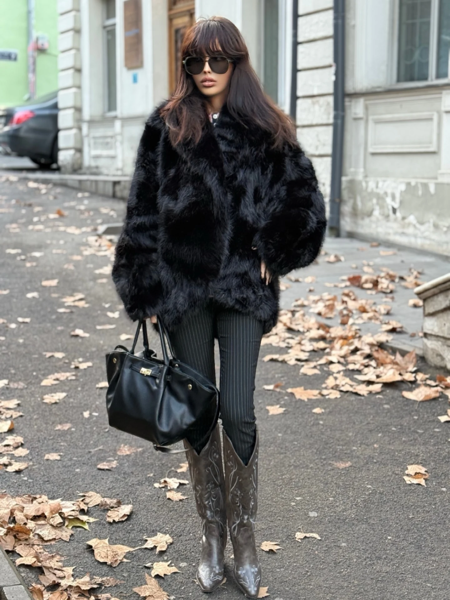 Woman wearing a black faux fur coat, pinstripe trousers, embroidered brown cowboy boots, and dark sunglasses.