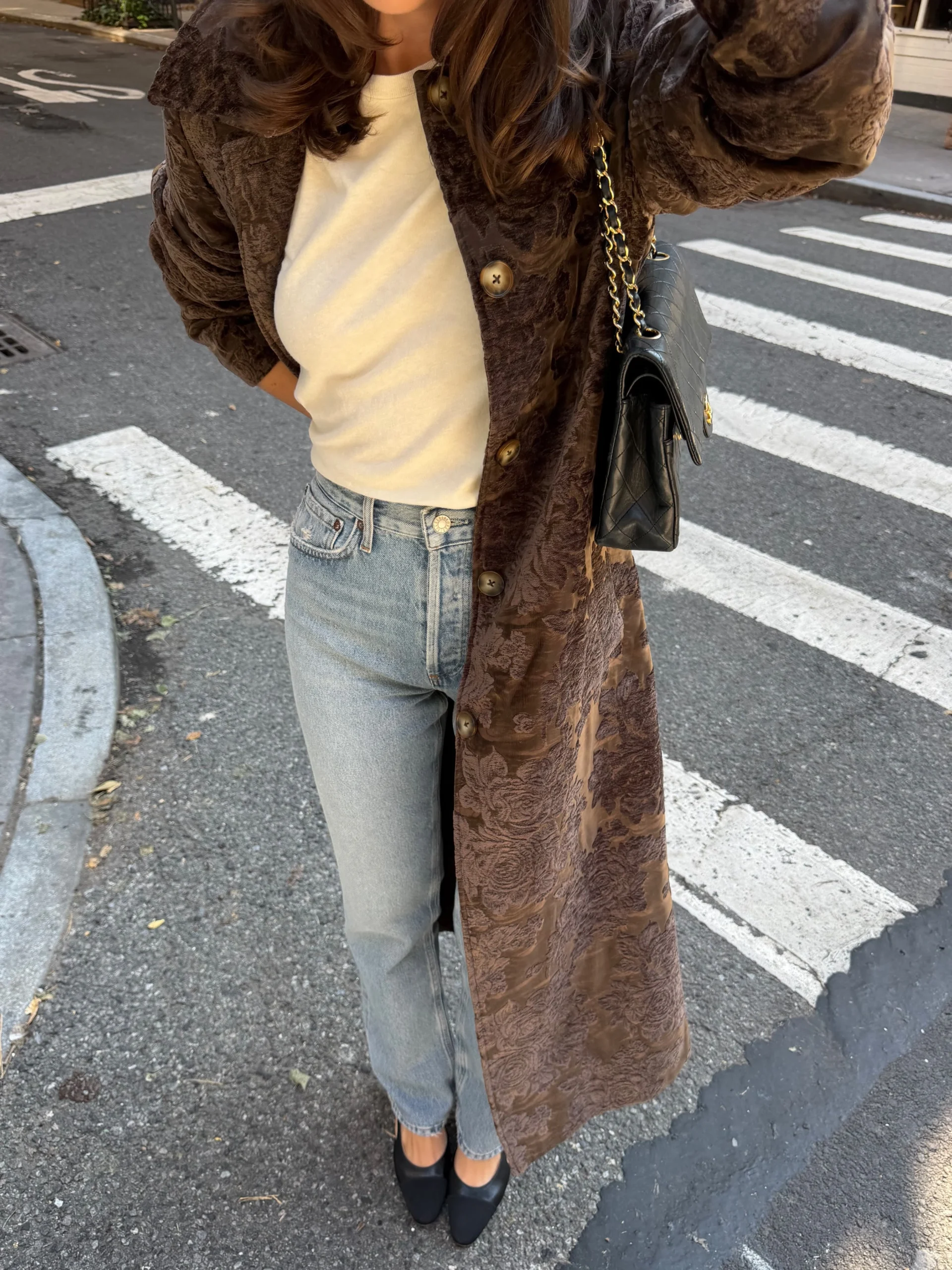 Woman wearing a long brown patterned coat, cream top, light-wash jeans, and black heeled shoes.