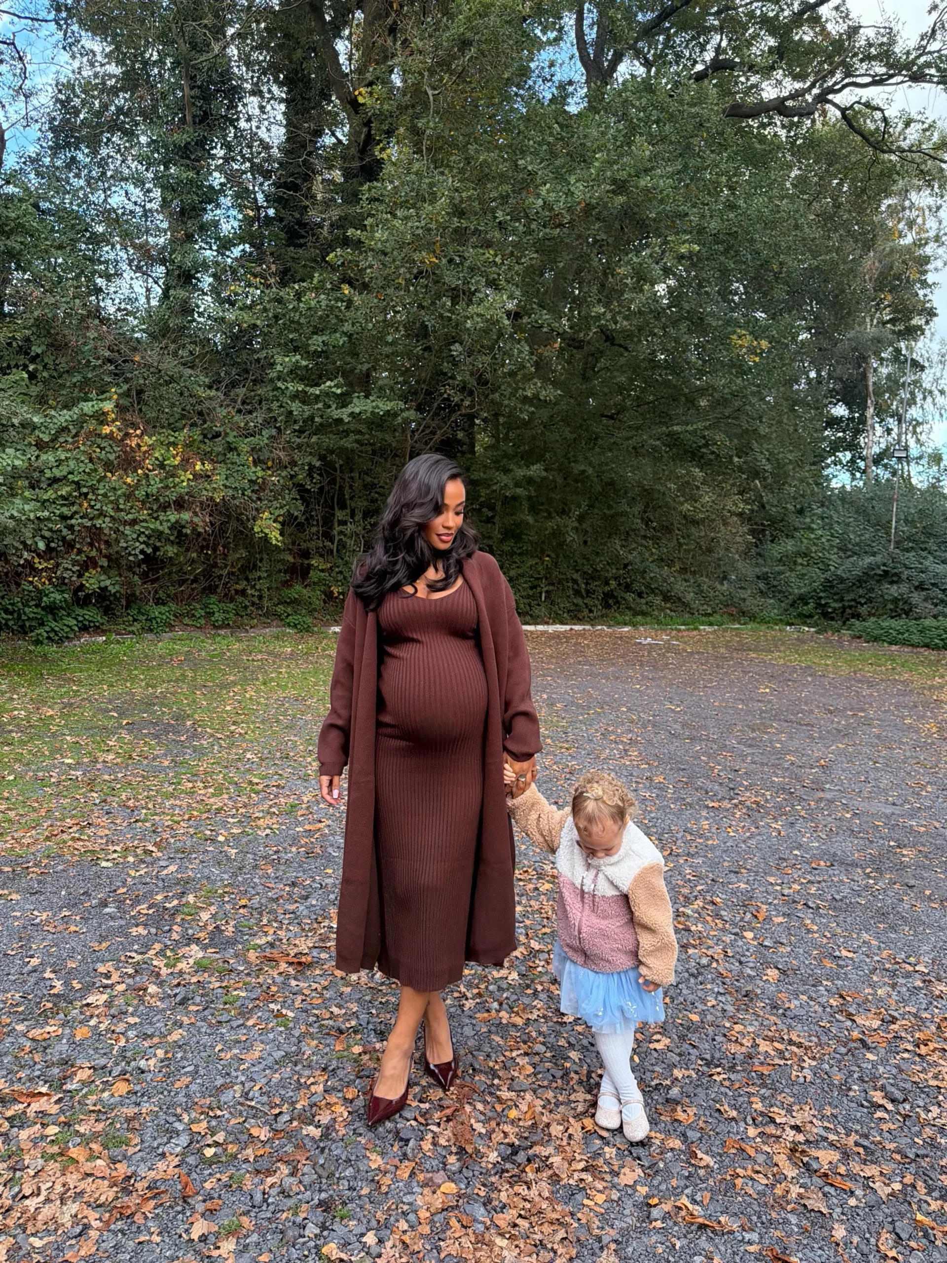 Pregnant woman in a brown ribbed dress and long coat with dark heels beside a child.