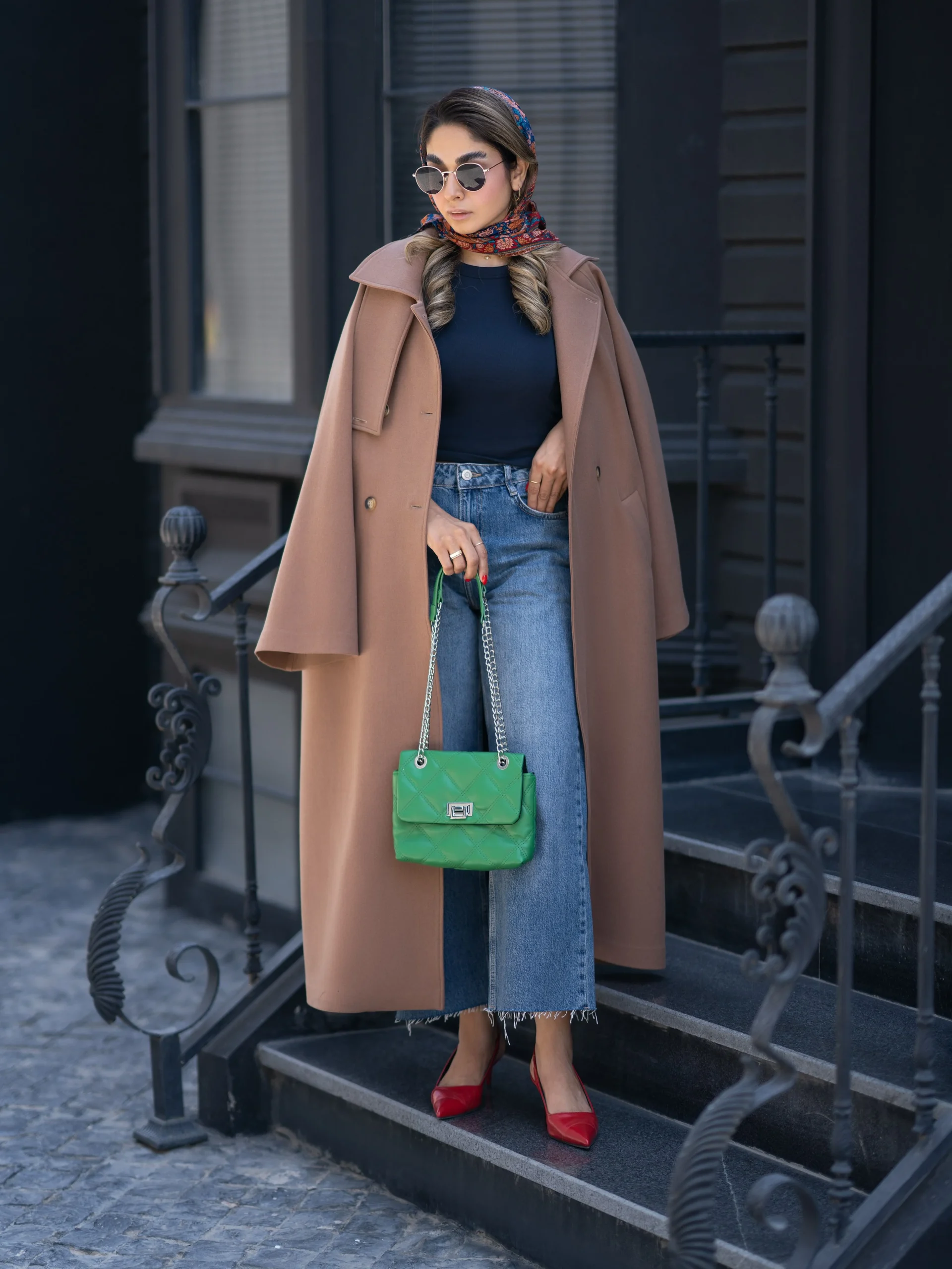 Long camel coat over cropped jeans styled with red heels, a green bag, and patterned scarf.