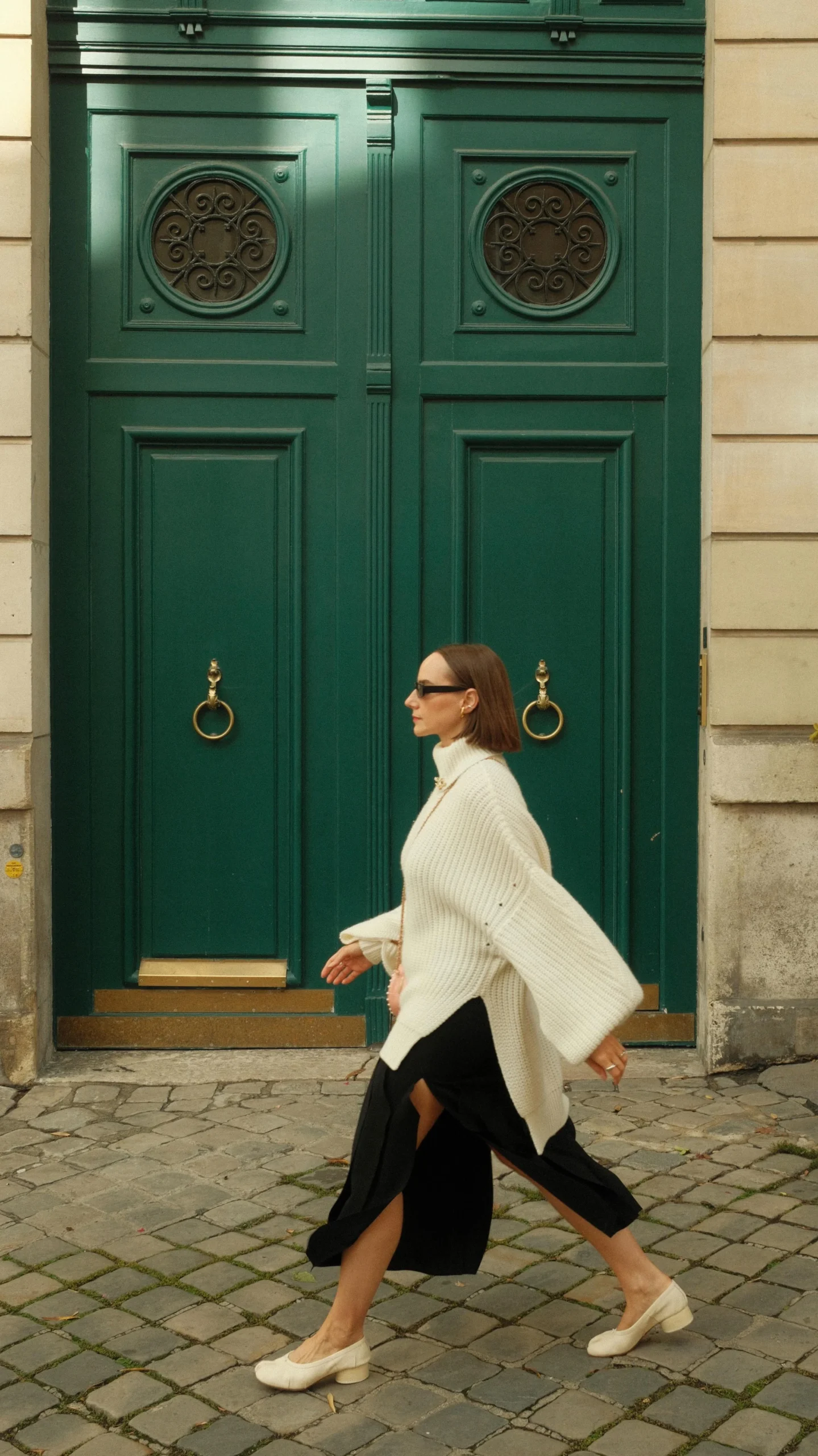 Person in cream oversized turtleneck, black cropped trousers, and beige flats walks past a dark green door.