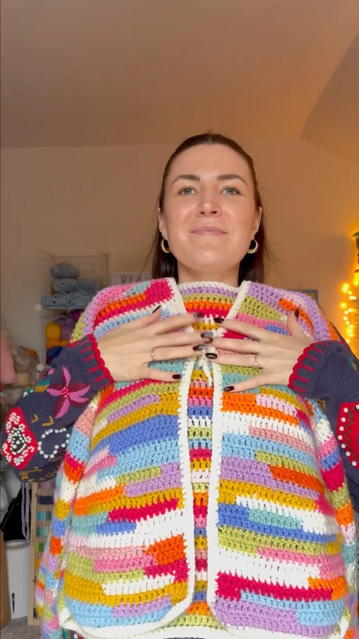 Loose open-front crochet cardigan with rainbow stripes, embroidered navy sleeves, and gold hoop earrings.