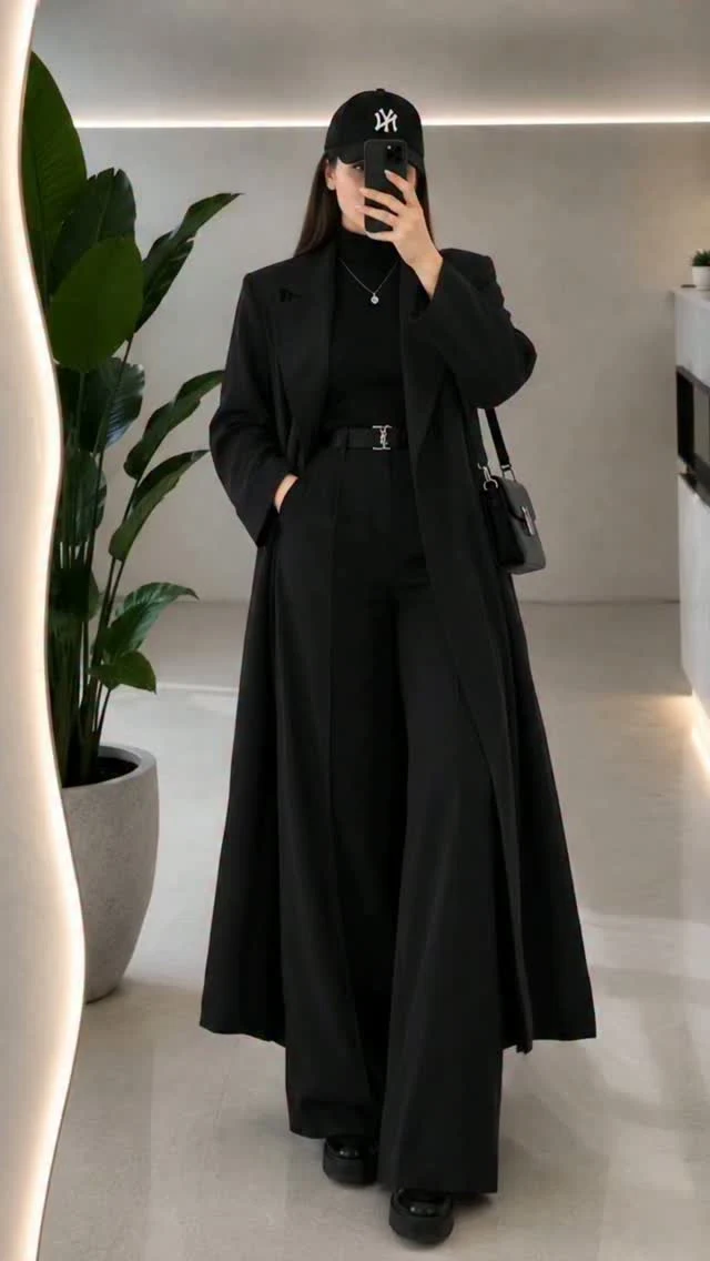 Person wearing an all-black outfit with oversized coat, wide-leg trousers, platform shoes, and baseball cap.